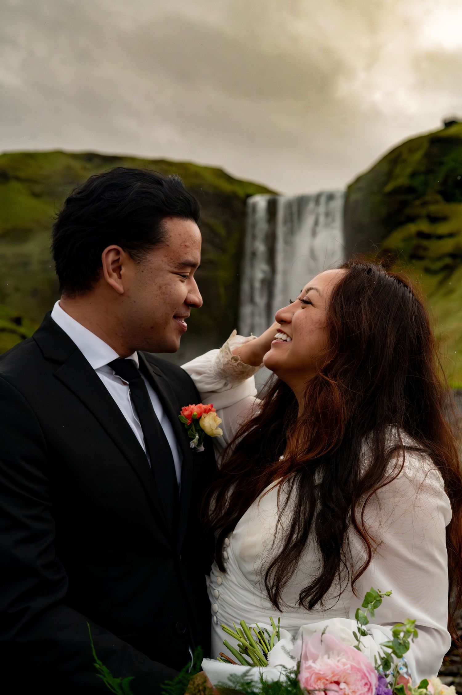 Couple sharing a romantic moment in front of Skogafoss waterfall in Iceland, bride holding colorful bouquet.