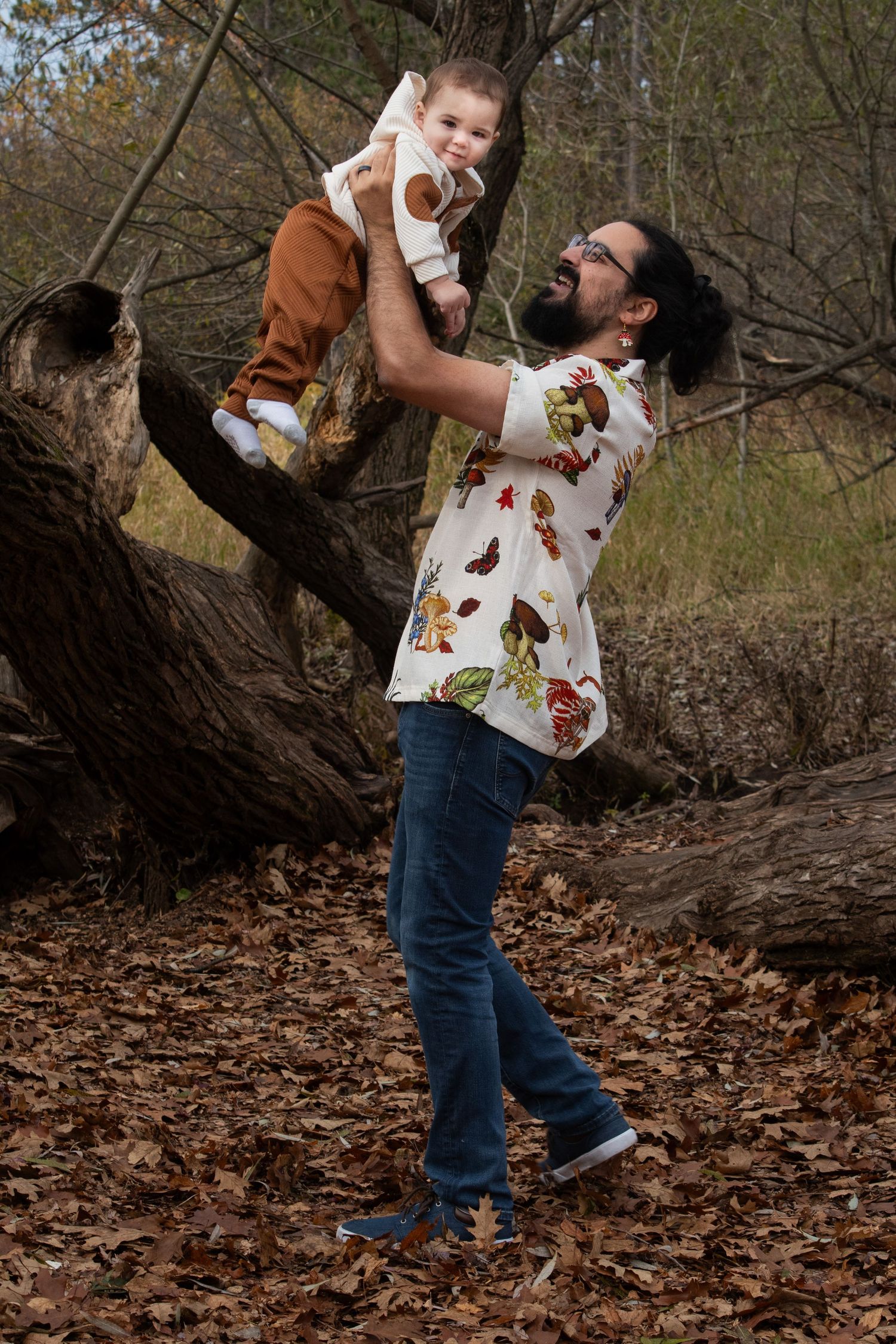Father in floral shirt lifting laughing baby overhead among fallen autumn leaves.