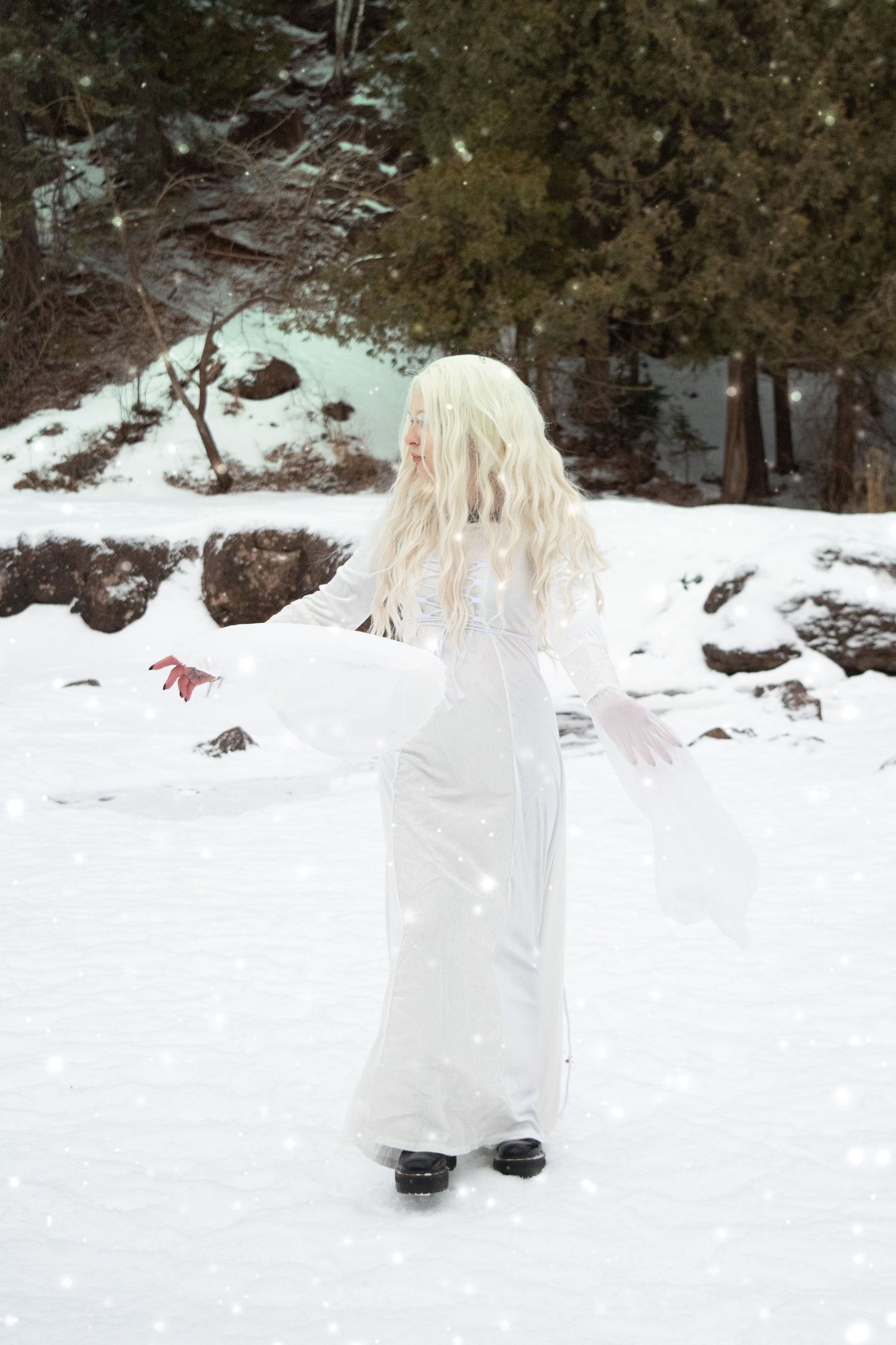 Blonde woman in elegant white gown standing in snowy forest during snowfall.