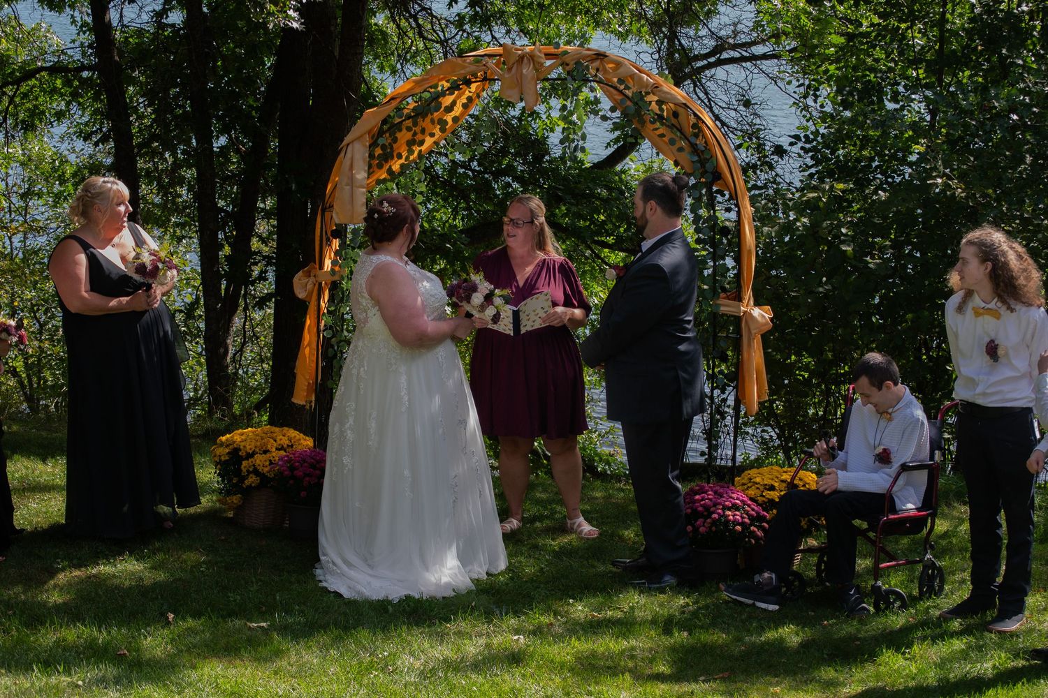 Outdoor wedding ceremony under floral arch with bride, groom, officiant, and guests on green lawn.