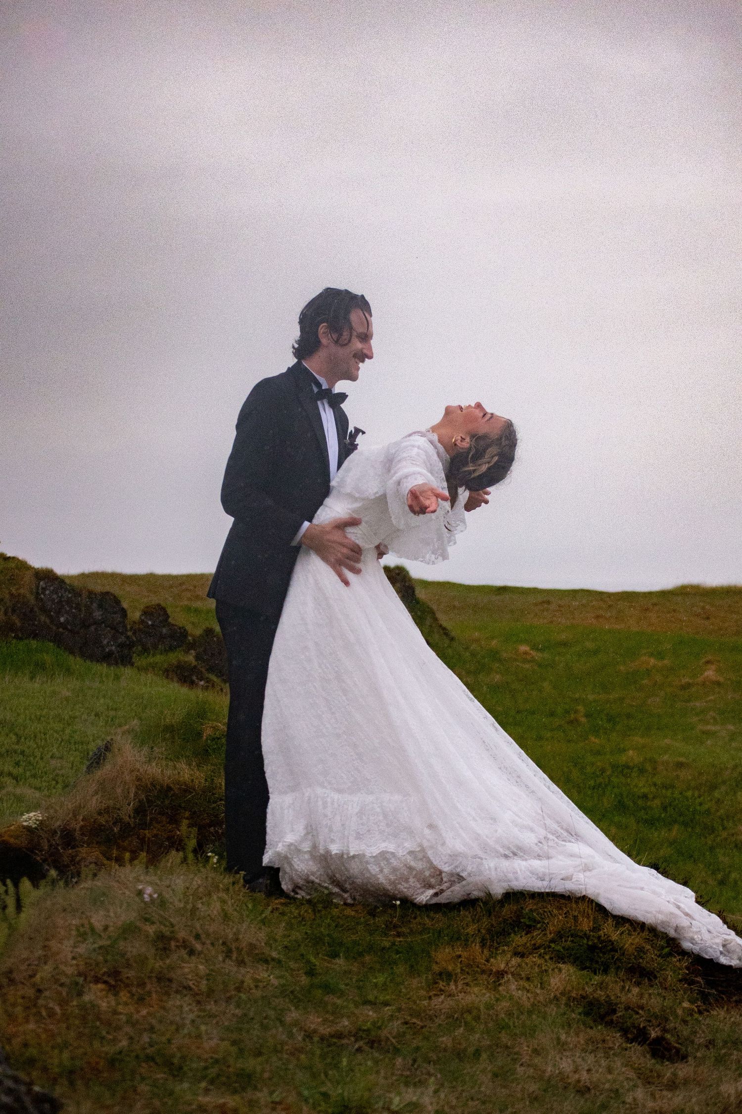 Bride and groom sharing joyful dance on misty hillside, her white lace gown flowing in wind during outdoor wedding shoot.