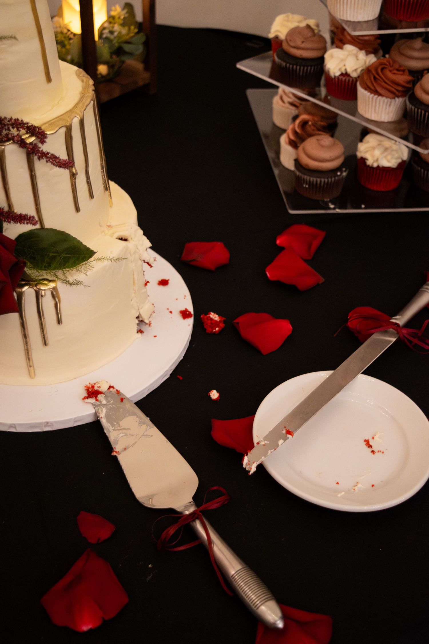 Wedding cake with red rose petals and serving utensils on a black table with cupcakes in the background.