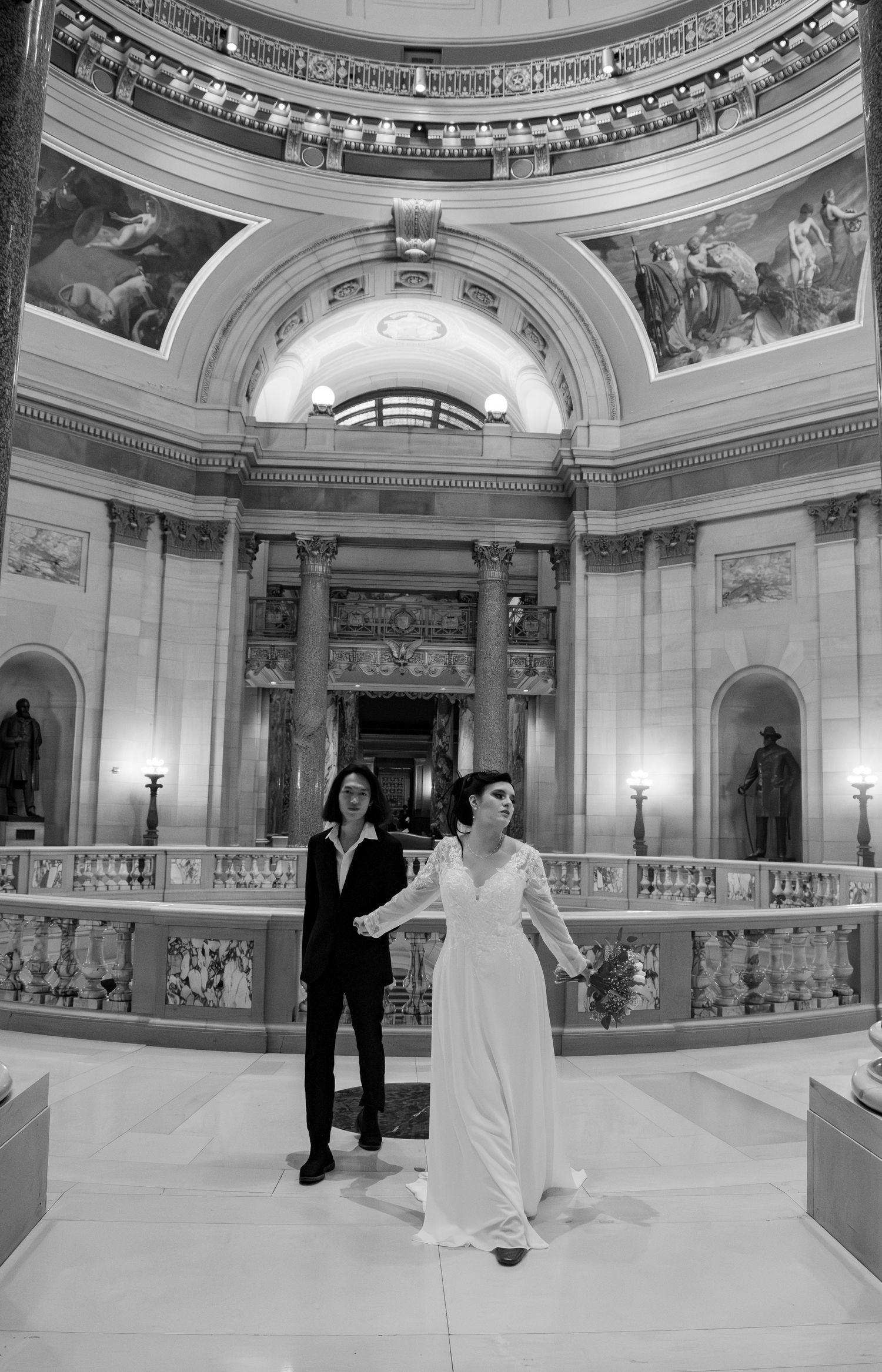 Black and white photo of newlywed couple holding hands in grand capitol rotunda with ornate architecture.