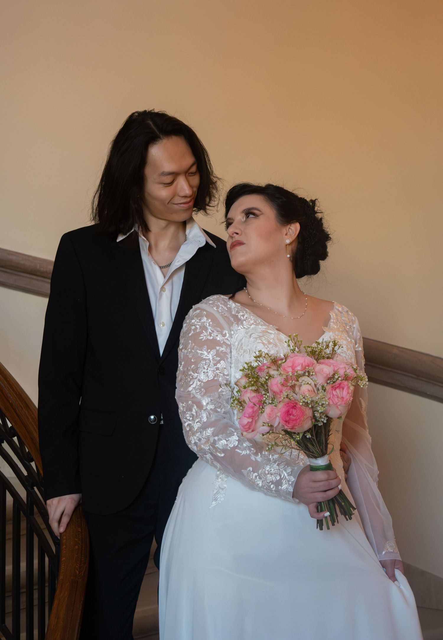 Bride in lace gown holding pink bouquet gazing at long-haired groom on staircase.