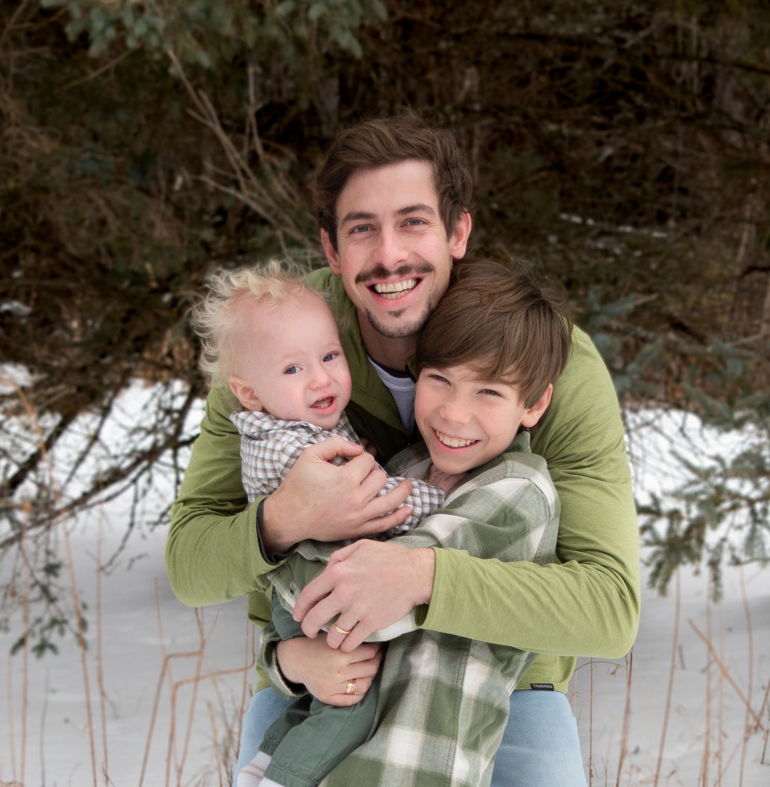 Father in green pullover hugging two sons tightly in snowy winter outdoor portrait.