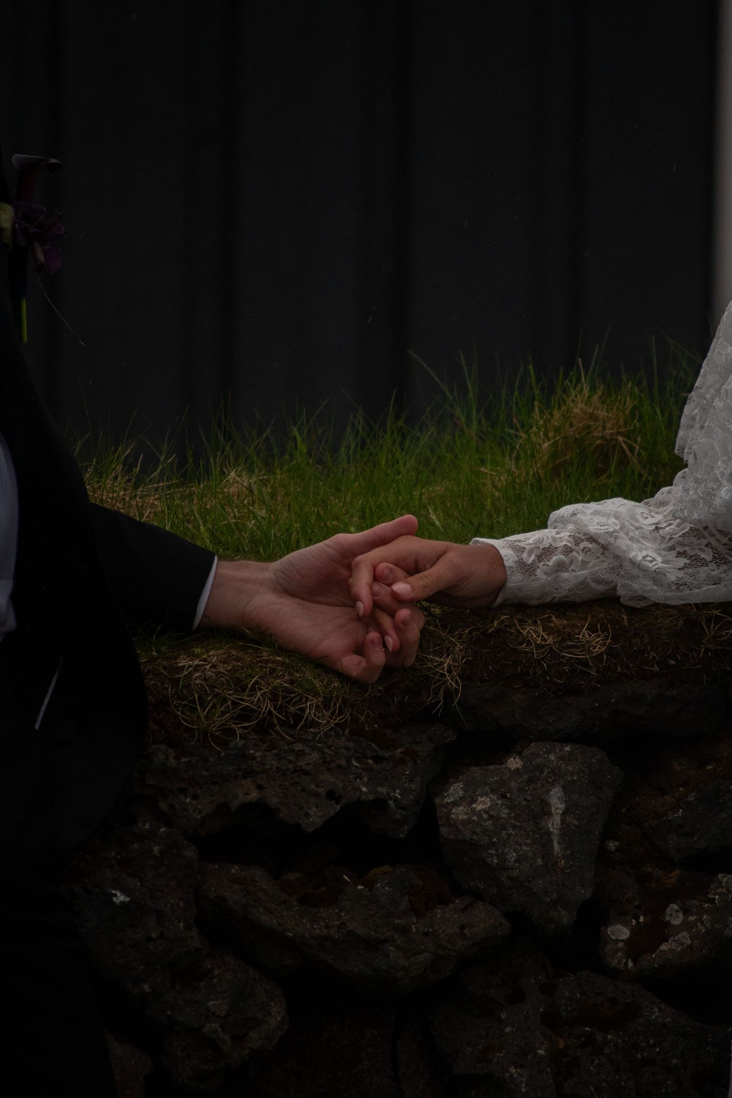 Bride and groom holding hands near a rustic stone wall with green grass backdrop during wedding ceremony.