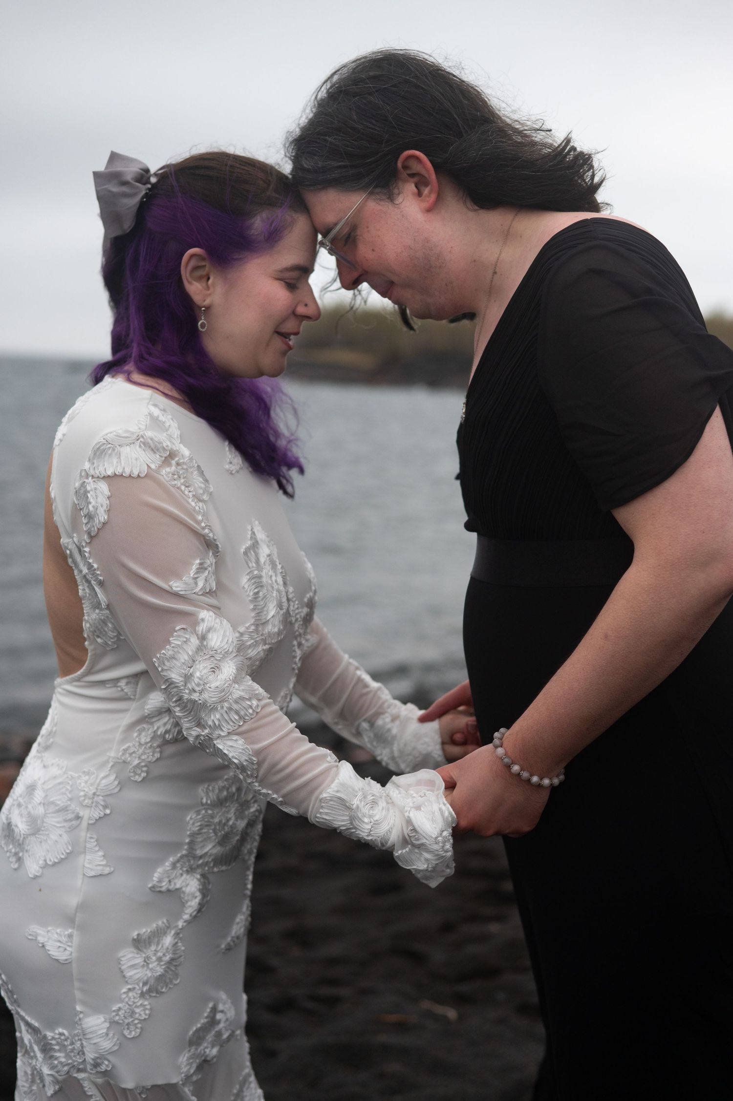 Two women in wedding attire hold hands and touch foreheads by the ocean, one in white lace gown with purple hair.