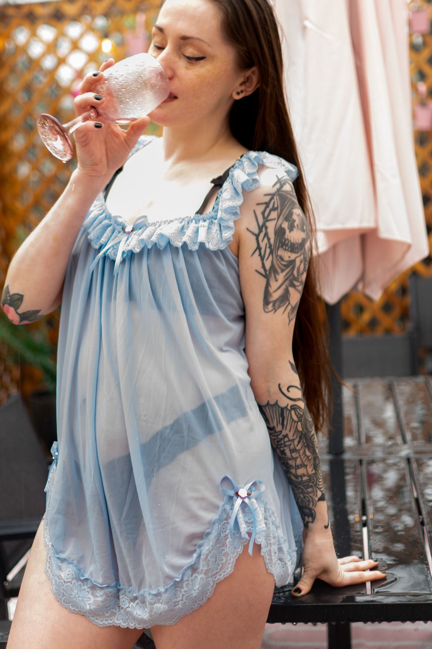 Tattooed woman in blue lace babydoll dress sipping from a pink cocktail glass outdoors.