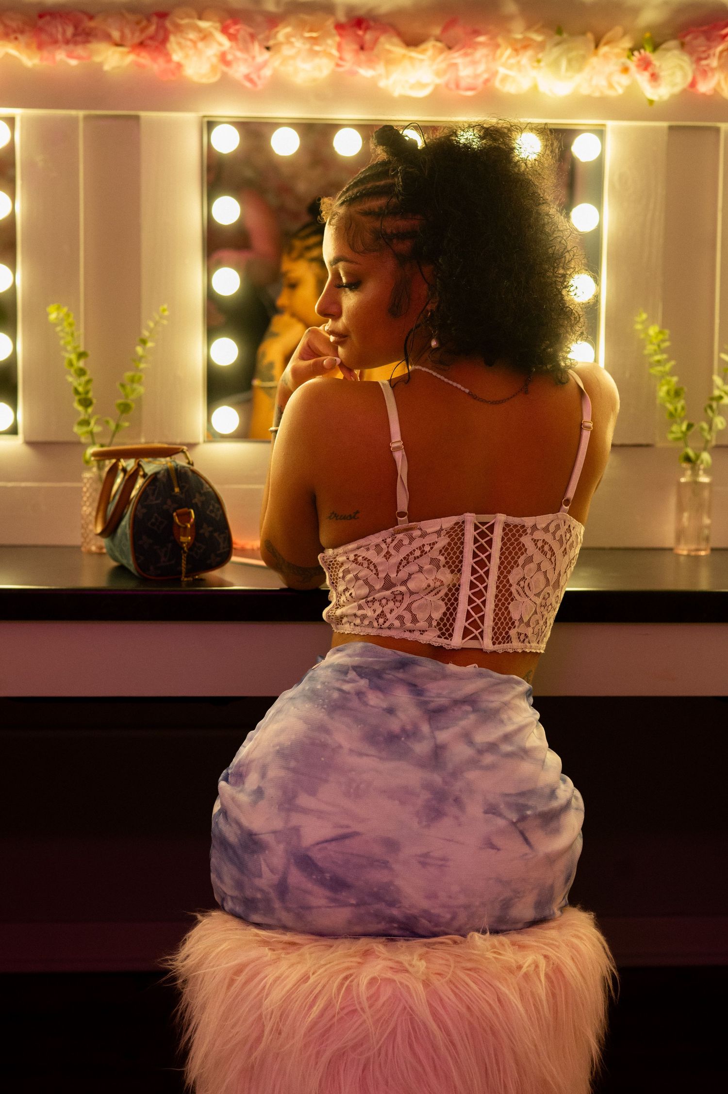 A young woman with curly hair sits at a vanity mirror wearing a white lace crop top and tie-dye skirt.