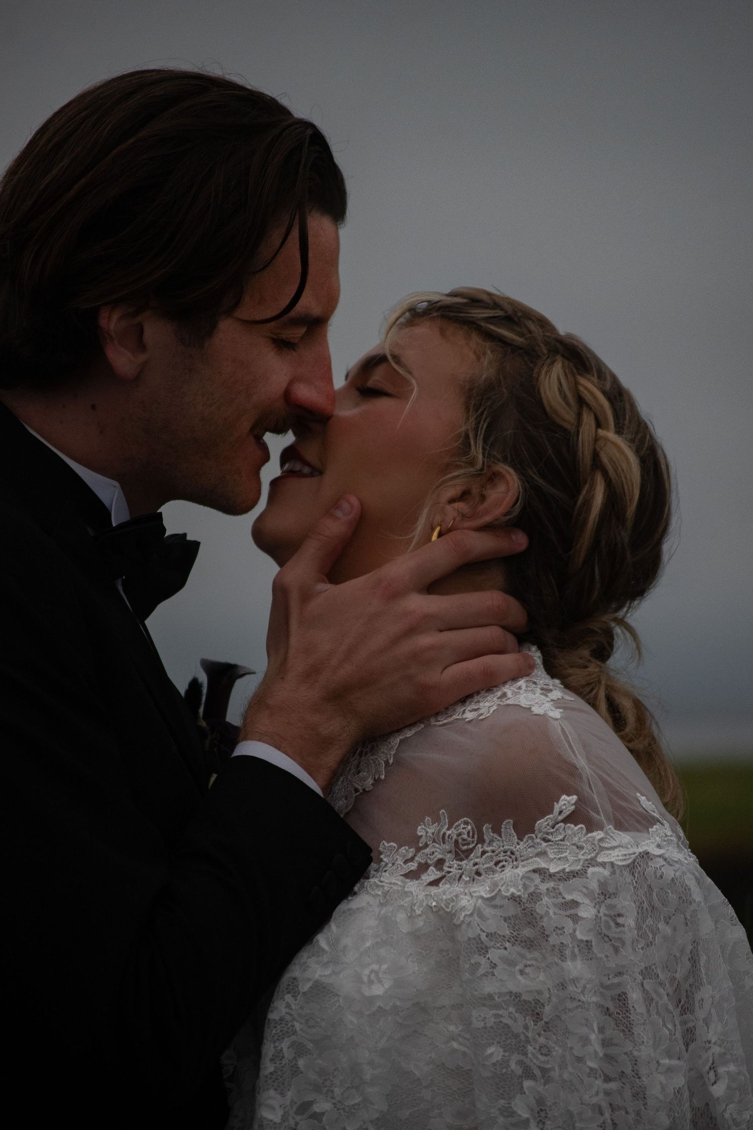A bride and groom share a romantic almost-kiss, the groom tenderly holding her face, both dressed in elegant wedding attire outdoors.