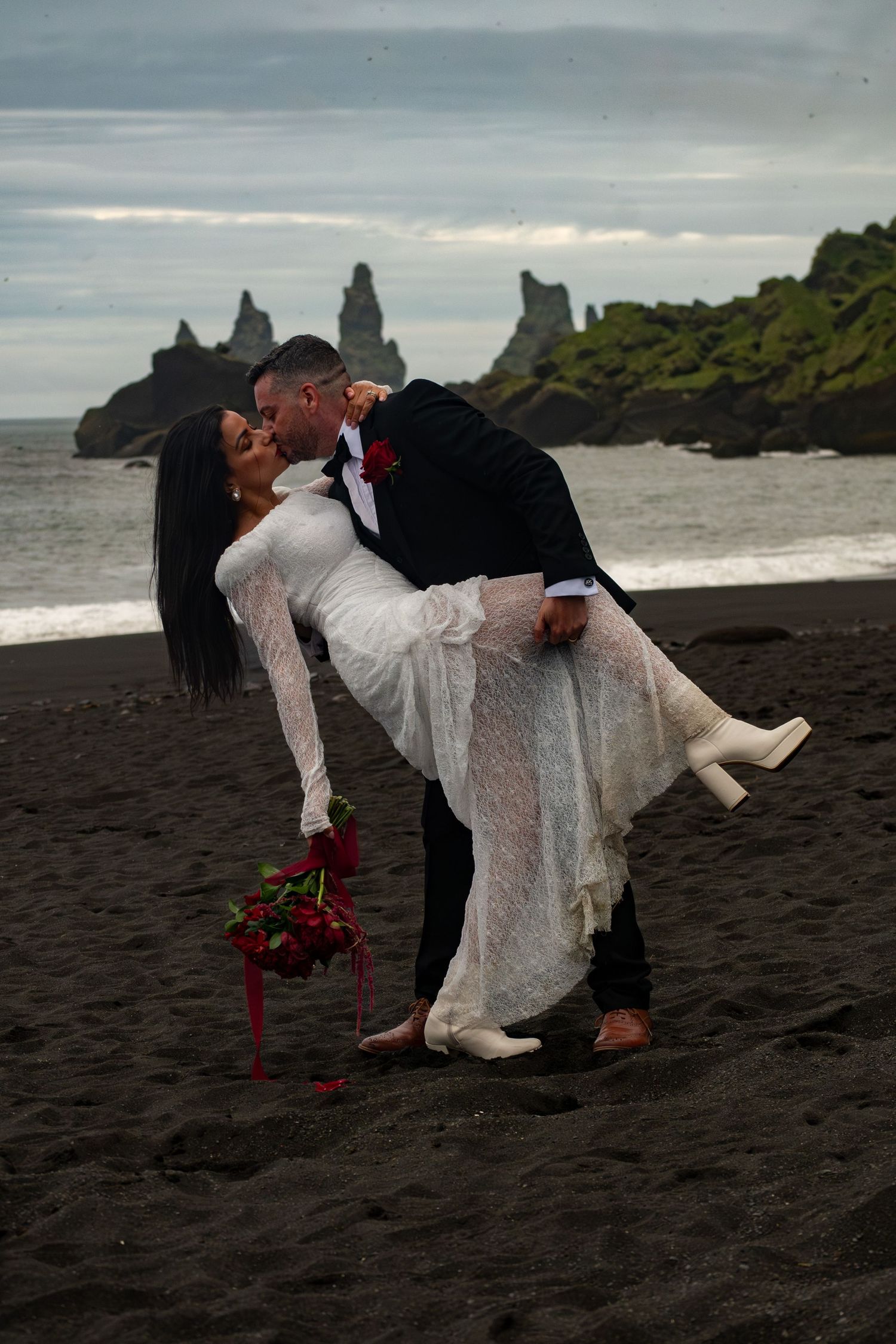 Couple sharing a romantic dip kiss on black sand beach in Iceland, bride in white dress holding red roses.