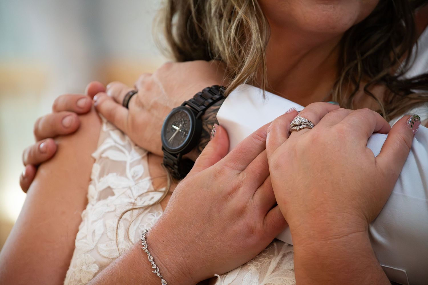 Close-up of bride and guest embracing, showcasing a black watch and diamond ring at a wedding.