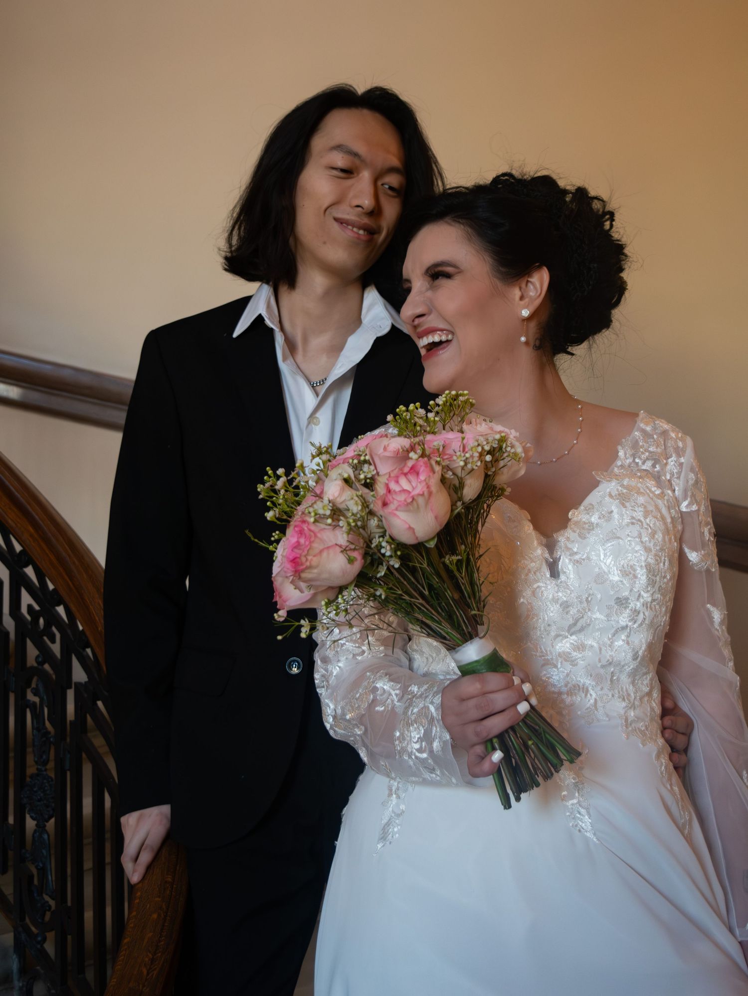 Joyful bride in lace wedding gown holding pink peony bouquet standing with groom on elegant staircase.