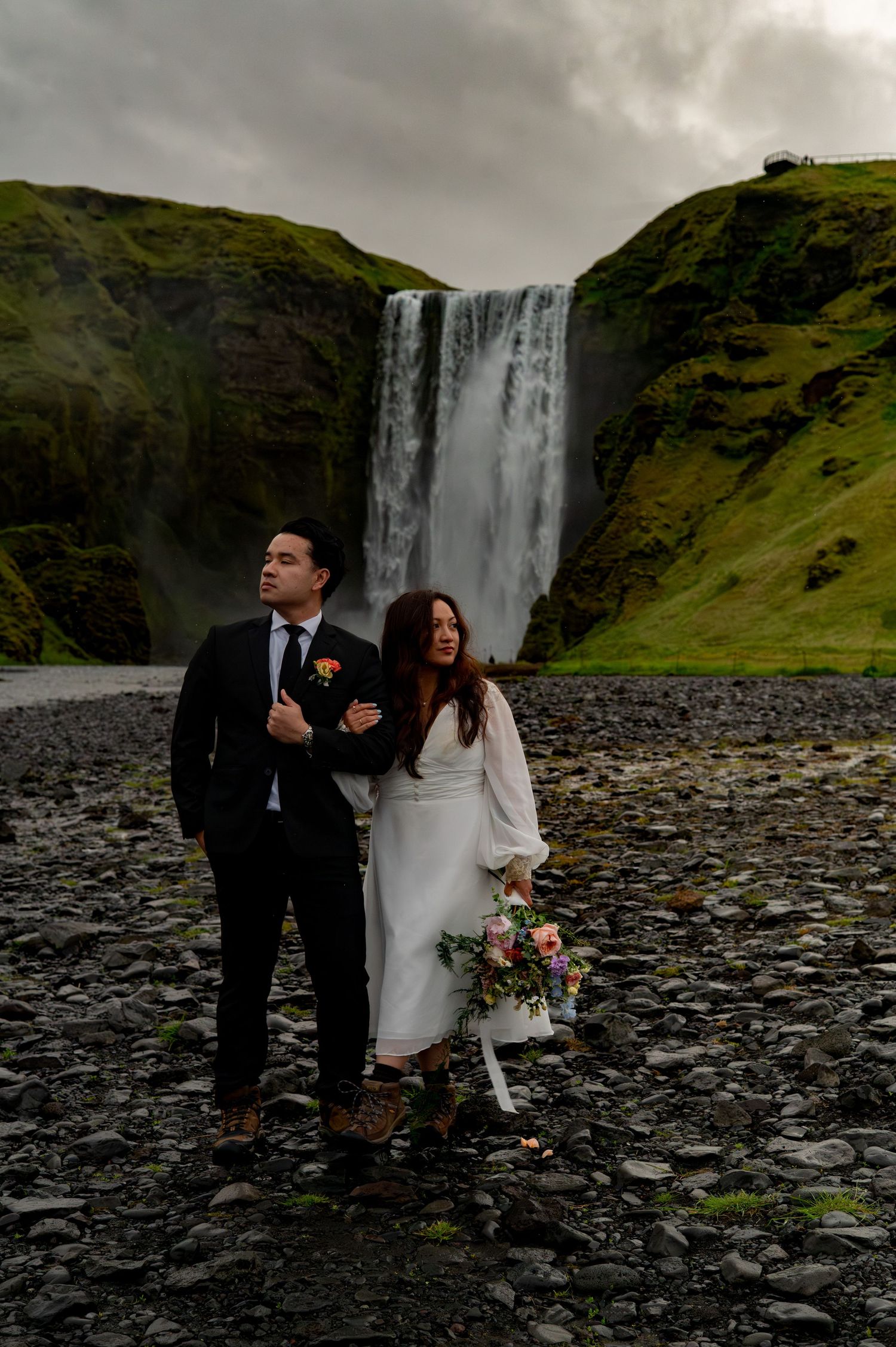 Couple eloping in front of Skogafoss waterfall in Iceland, bride in white dress holding bouquet, groom in dark suit.