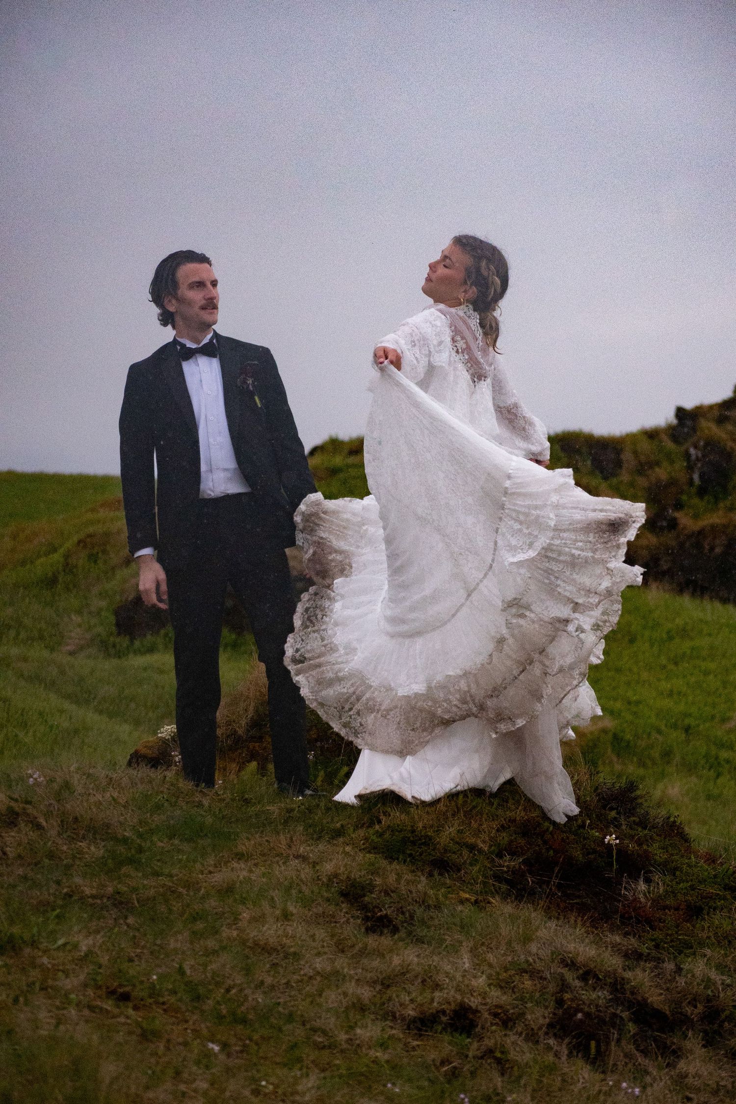 Groom in black tuxedo watches bride twirl her flowing lace wedding dress on a scenic grassy hillside.