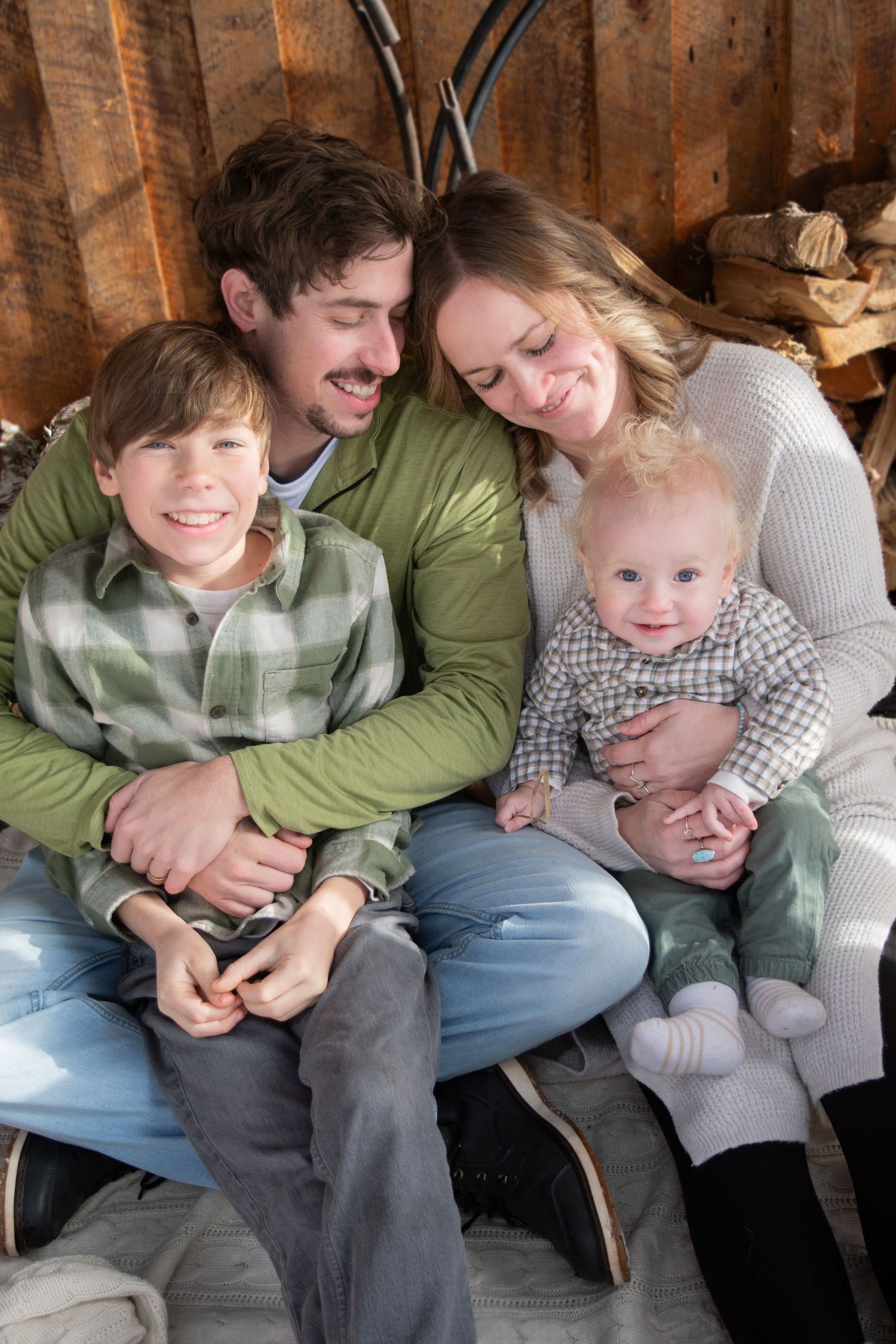 A happy family of four poses together in a rustic wooden setting, with two parents and their young son and baby.