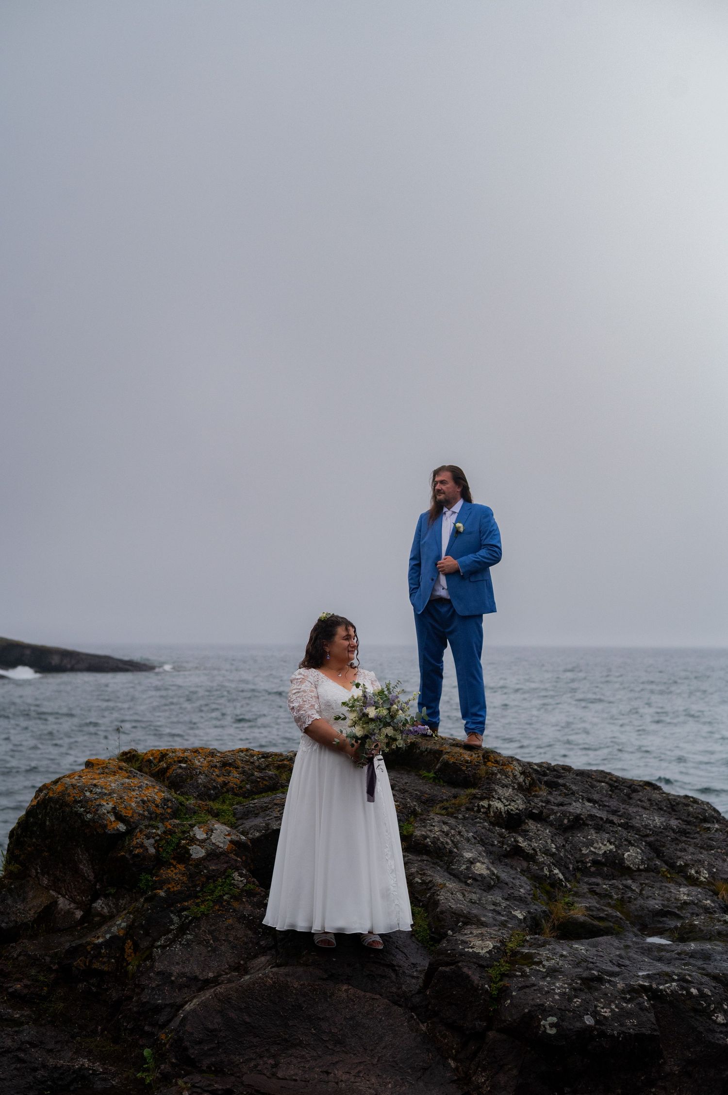 Bride in white gown and groom in blue suit pose on rocky coastal cliffs with ocean backdrop on their wedding day.
