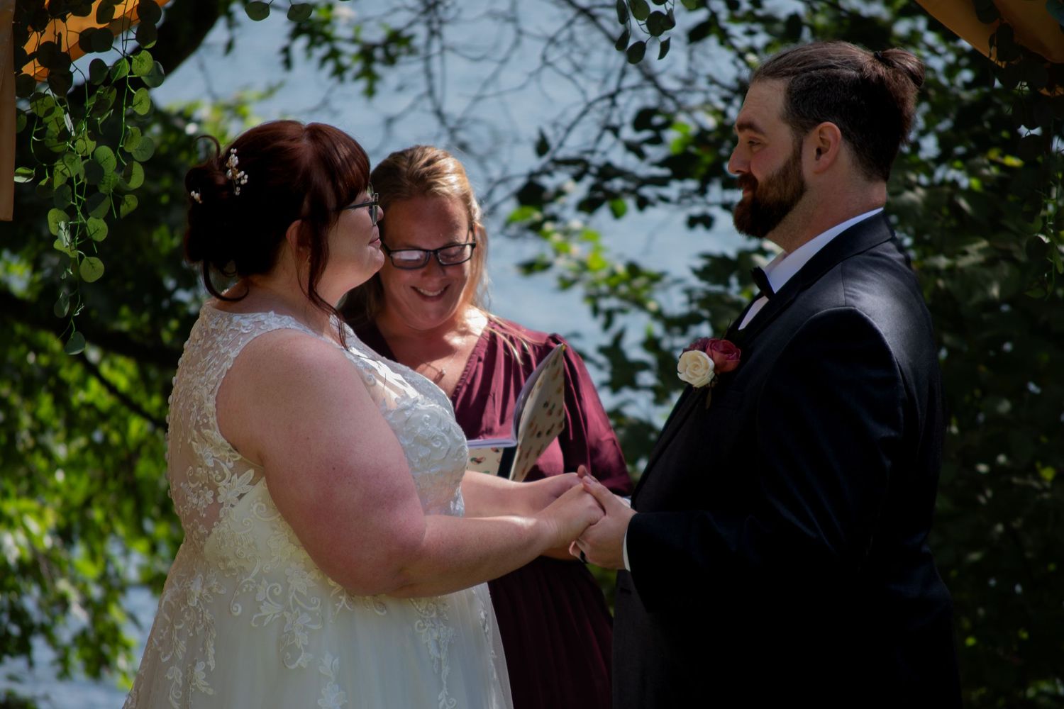 Outdoor wedding ceremony with bride in white gown, groom in suit, and officiant in burgundy dress exchanging vows under green trees.