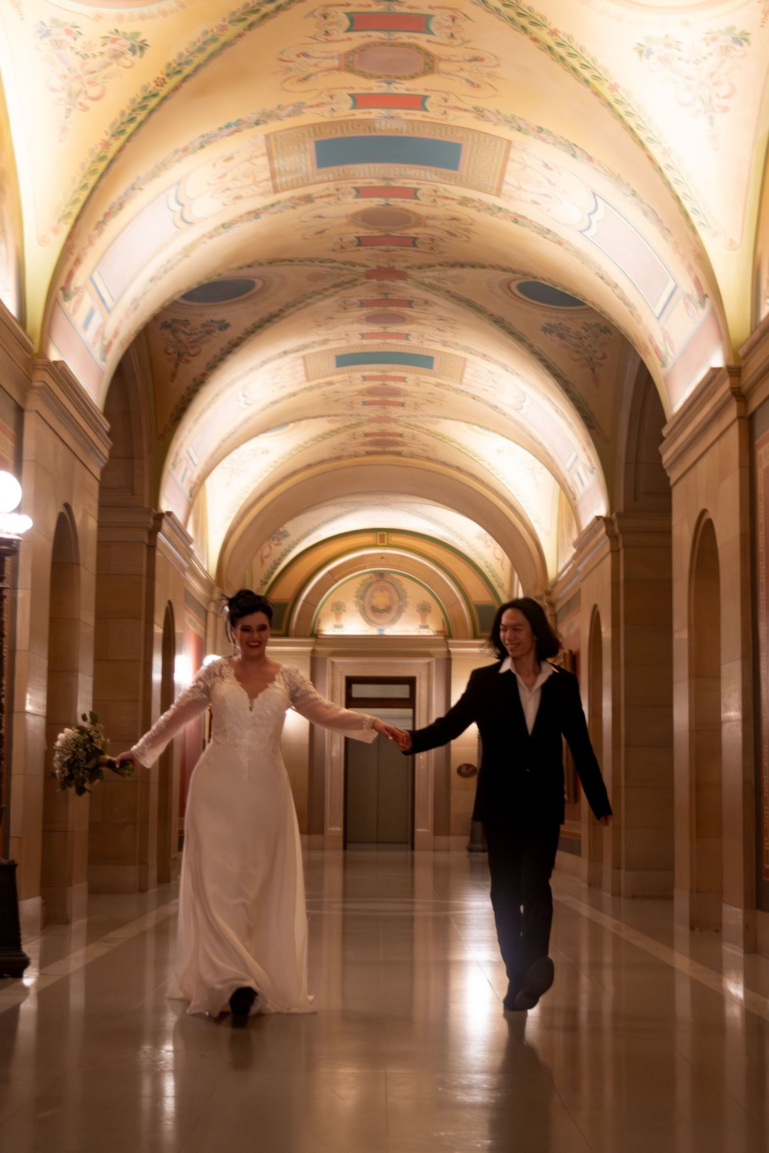 Bride and groom holding hands walking through ornate arched hallway of elegant venue.