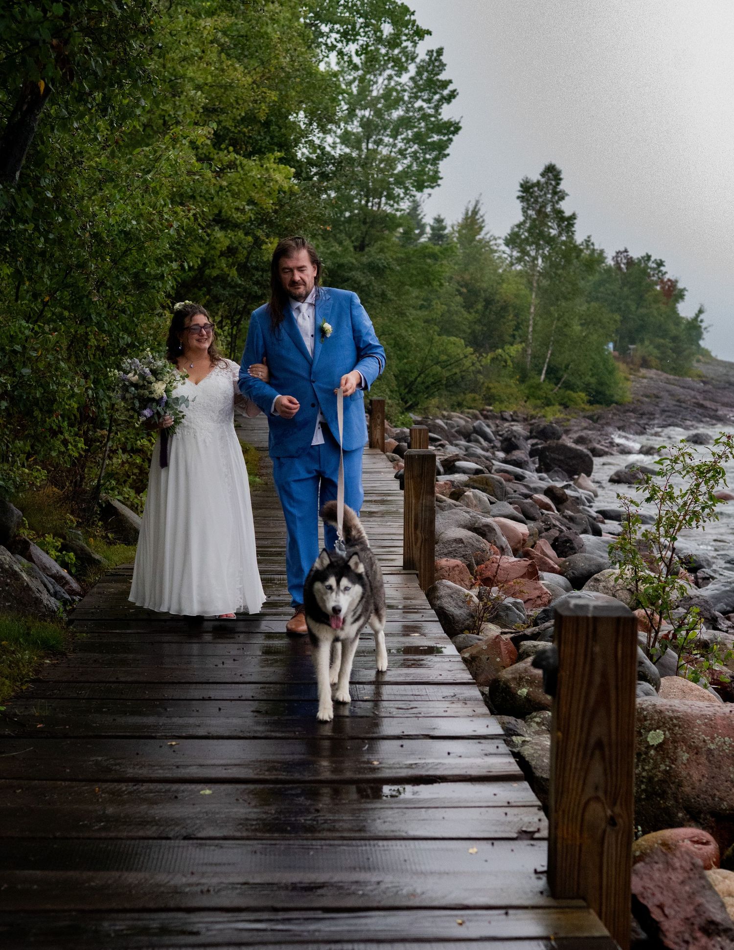 Bride and groom walking Husky dog on wooden boardwalk by rocky shoreline on their wedding day.