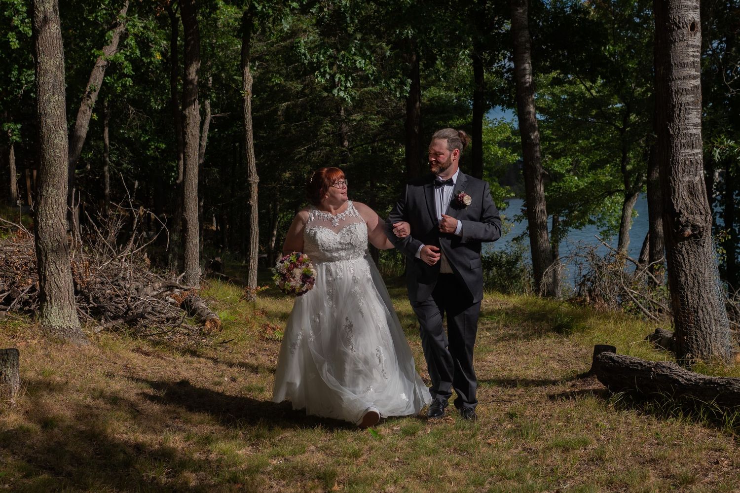 A bride and groom walk hand in hand through a sunlit forest, with the bride in a white gown and groom in a dark suit.