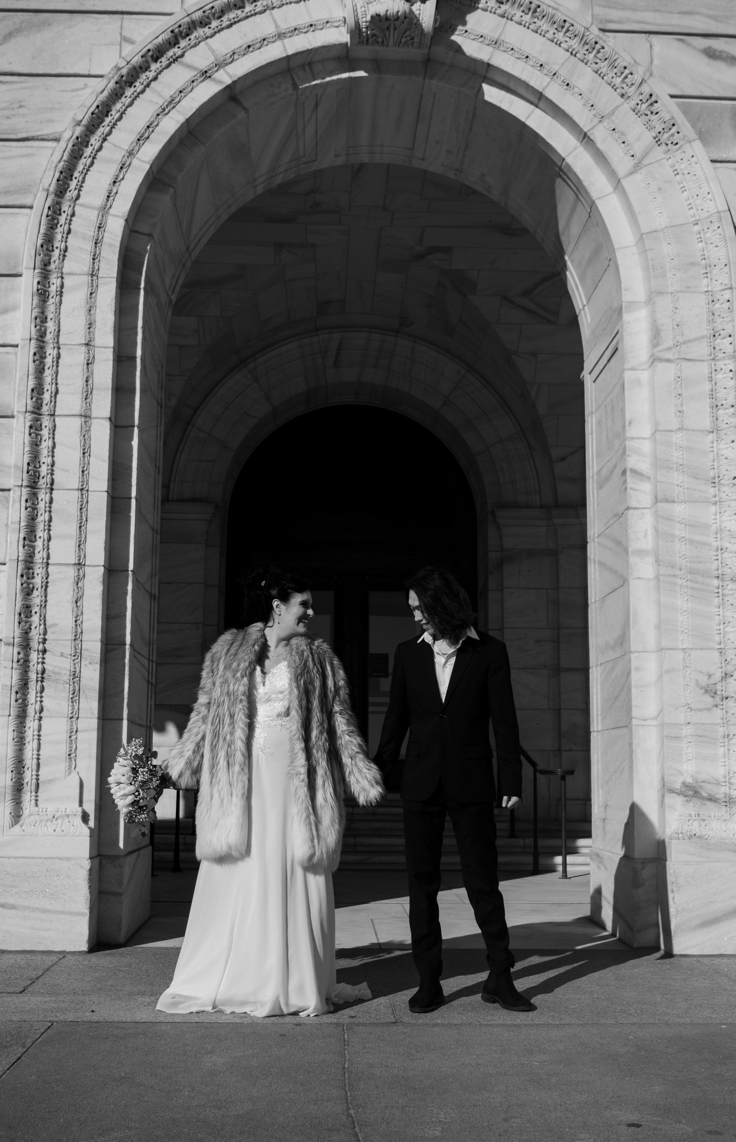 Black and white photo of newlywed couple holding hands under grand stone archway of historic building.