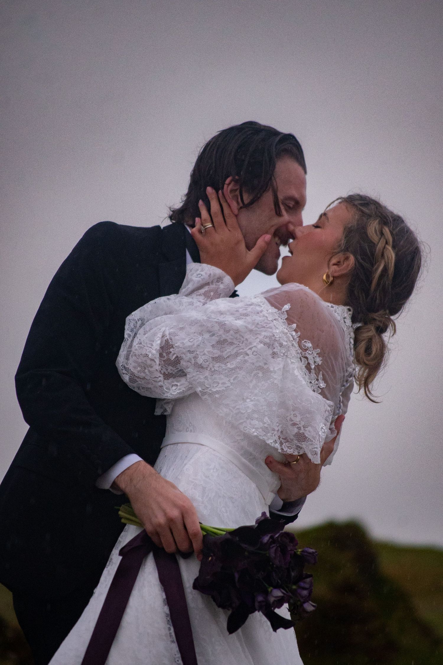 Bride and groom share a romantic kiss outdoors, bride holding dark purple floral bouquet on their wedding day.