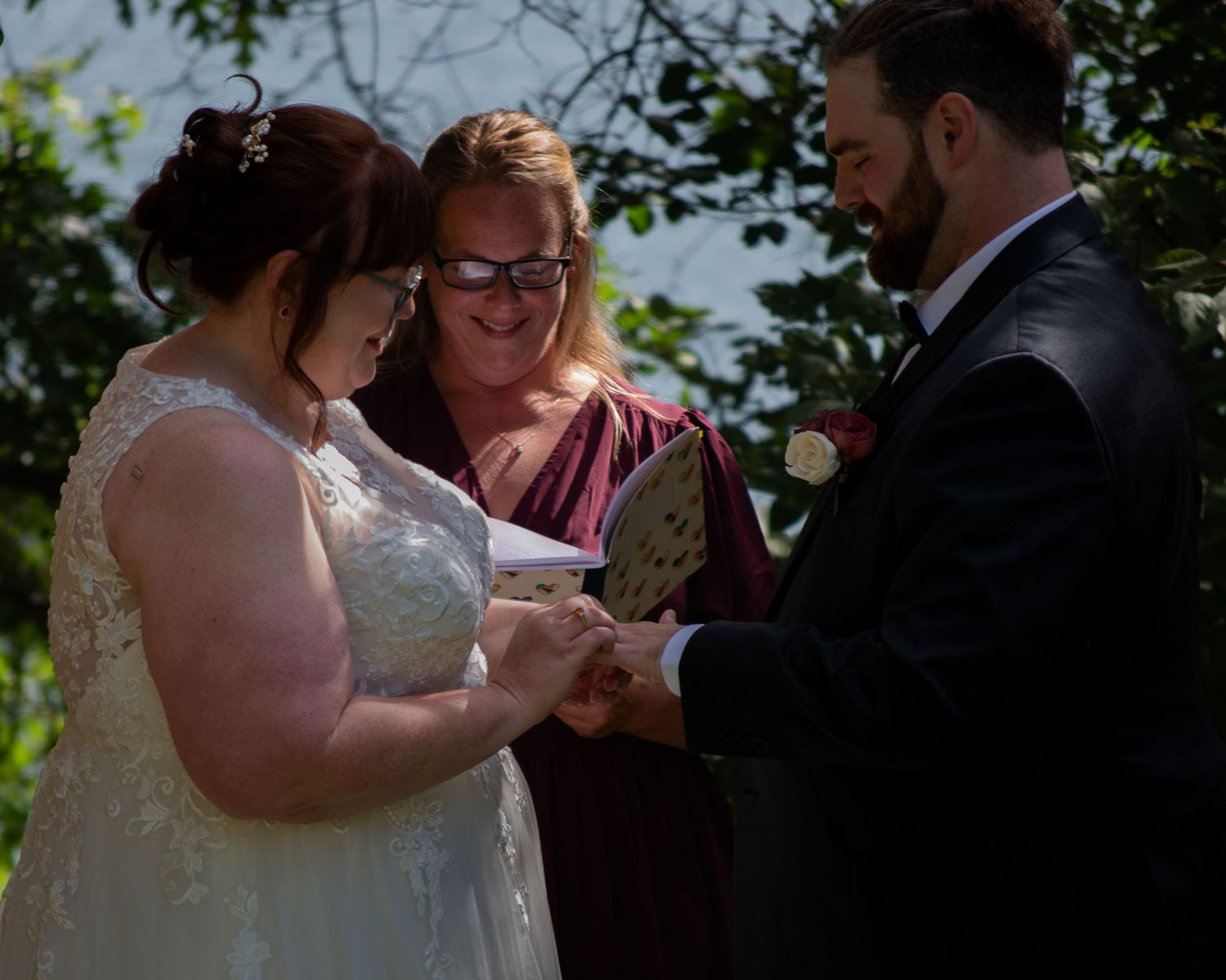 Bride and groom exchange rings during outdoor wedding ceremony with officiant reading from book under green trees.