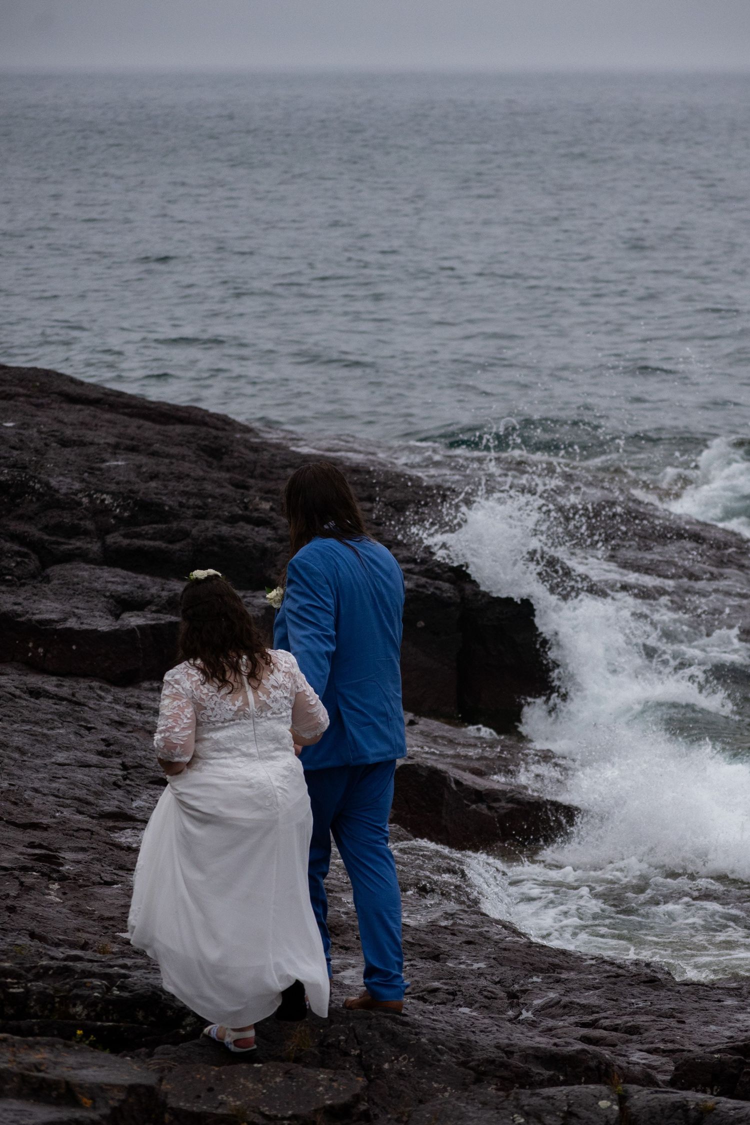 Bride in white dress and groom in blue suit stand on rocky coastal cliffs watching ocean waves crash below.