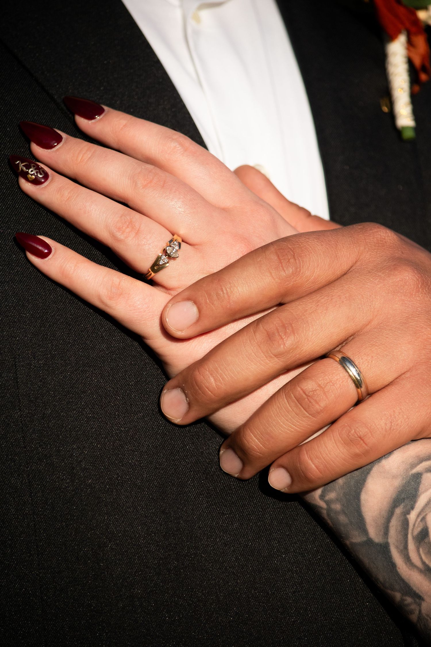 Close-up of bride and groom hands showing wedding rings, dark red nails and rose tattoo on wrist.