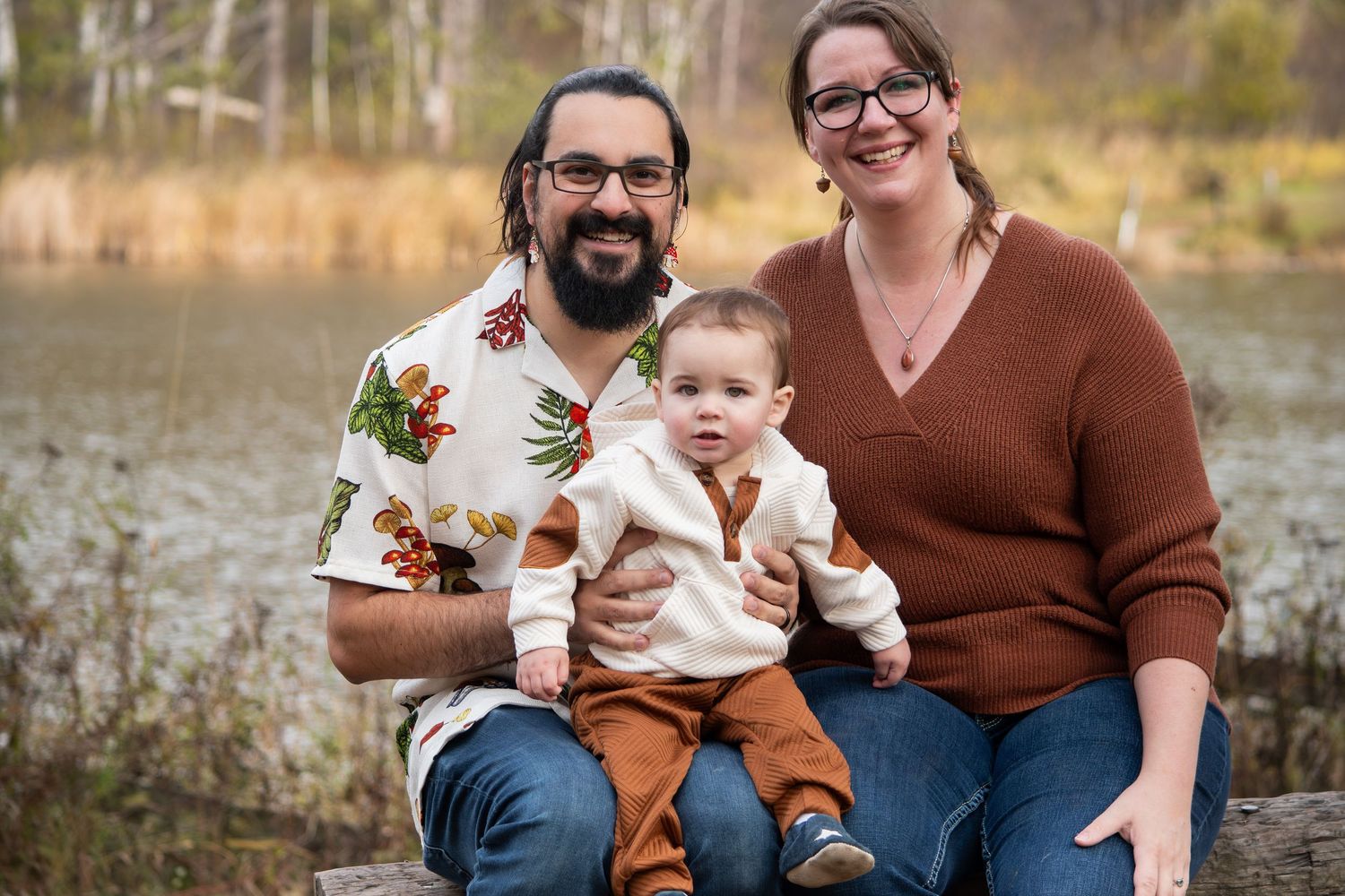 Smiling couple with baby sitting on log in autumn forest setting.