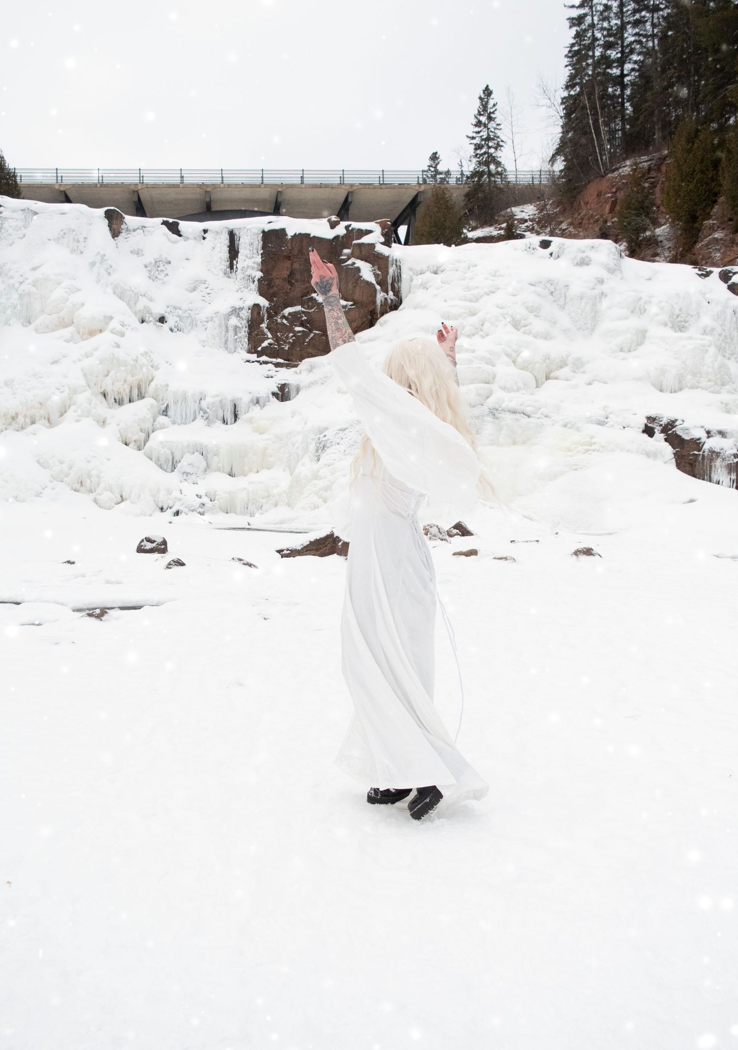 Woman in white dress and veil dancing in snowy winter landscape near frozen waterfall.