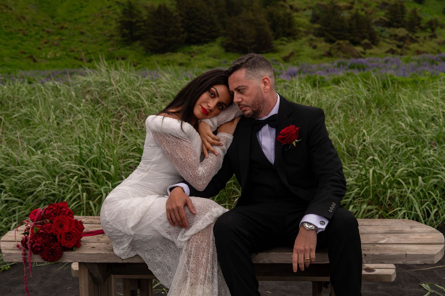 Bride in white lace gown and groom in black tuxedo sitting on bench with red bouquet, sharing romantic moment outdoors.