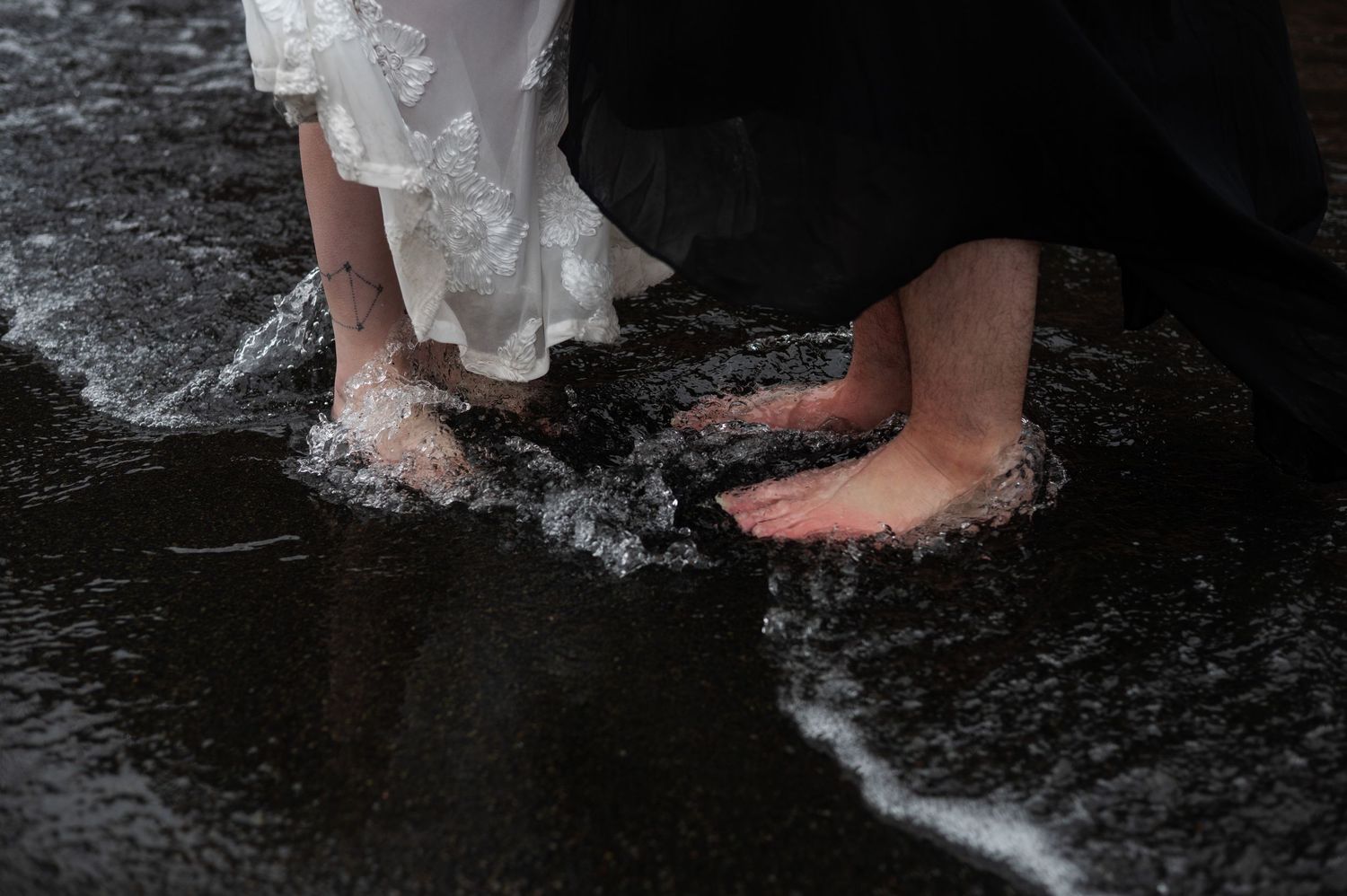 Two people standing barefoot in shallow ocean waves on a dark sandy beach, wearing formal white and black attire.