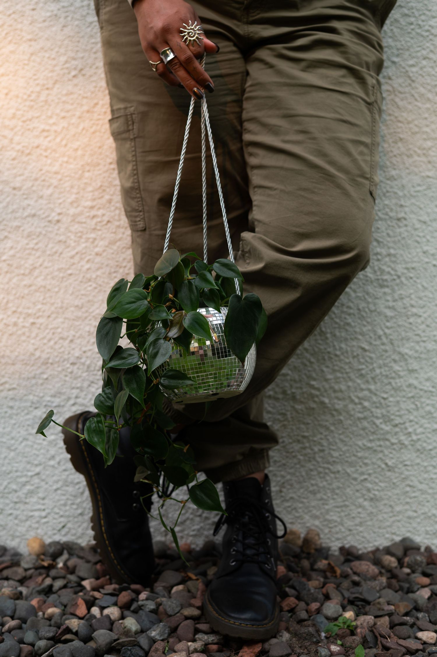 A person in olive cargo pants and black boots holds a disco ball planter with trailing green pothos vines.