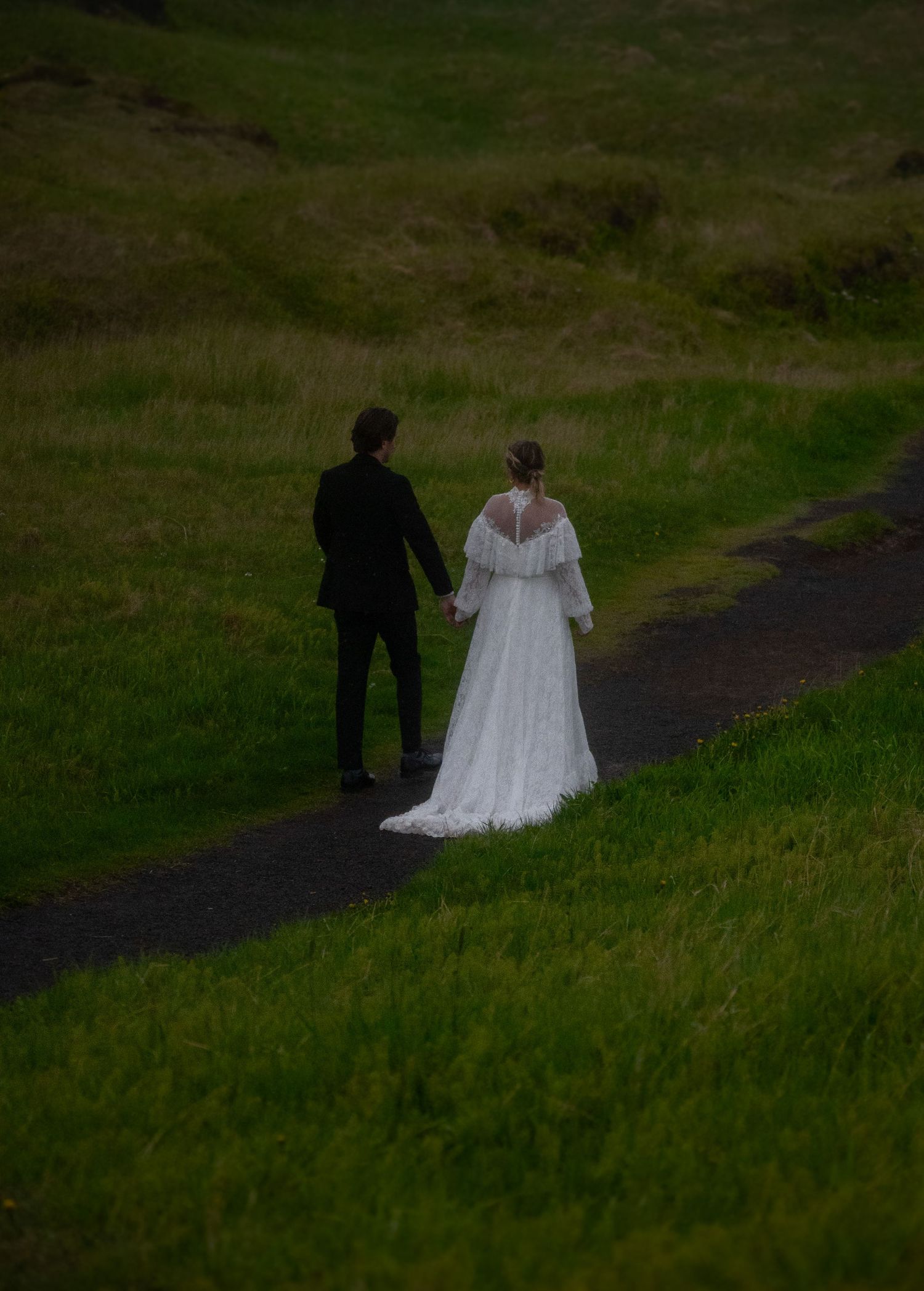 Bride and groom walk hand in hand along a winding path through lush green fields on their wedding day.