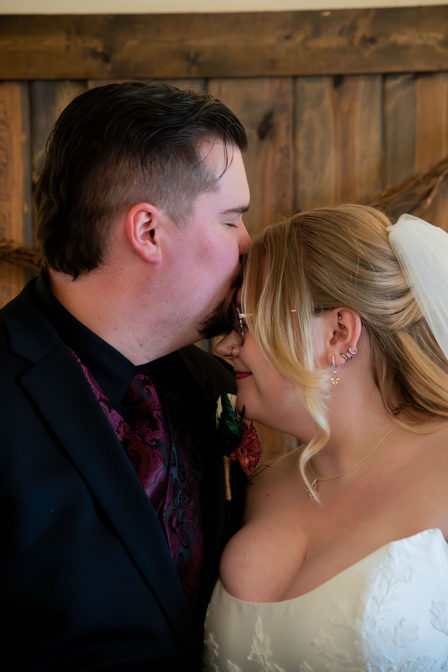 Groom in black suit with burgundy tie kisses bride in white lace wedding dress on forehead.