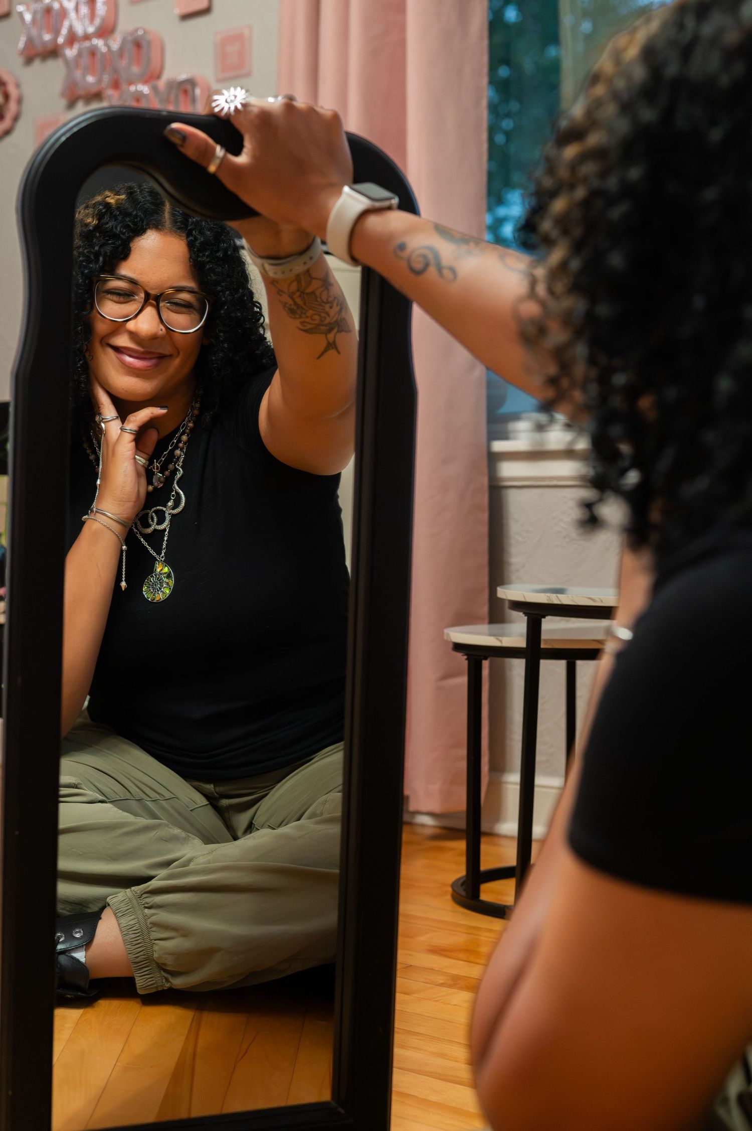 A tattooed woman with curly hair and glasses sits cross-legged, smiling at her reflection in a black mirror.