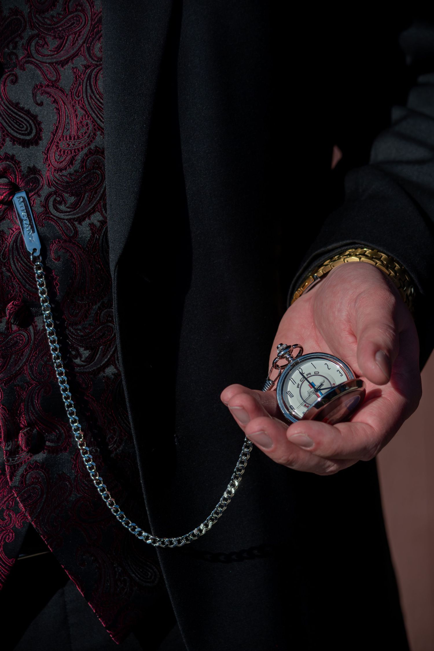 Groom holds vintage silver pocket watch on chain against burgundy paisley wedding suit jacket.