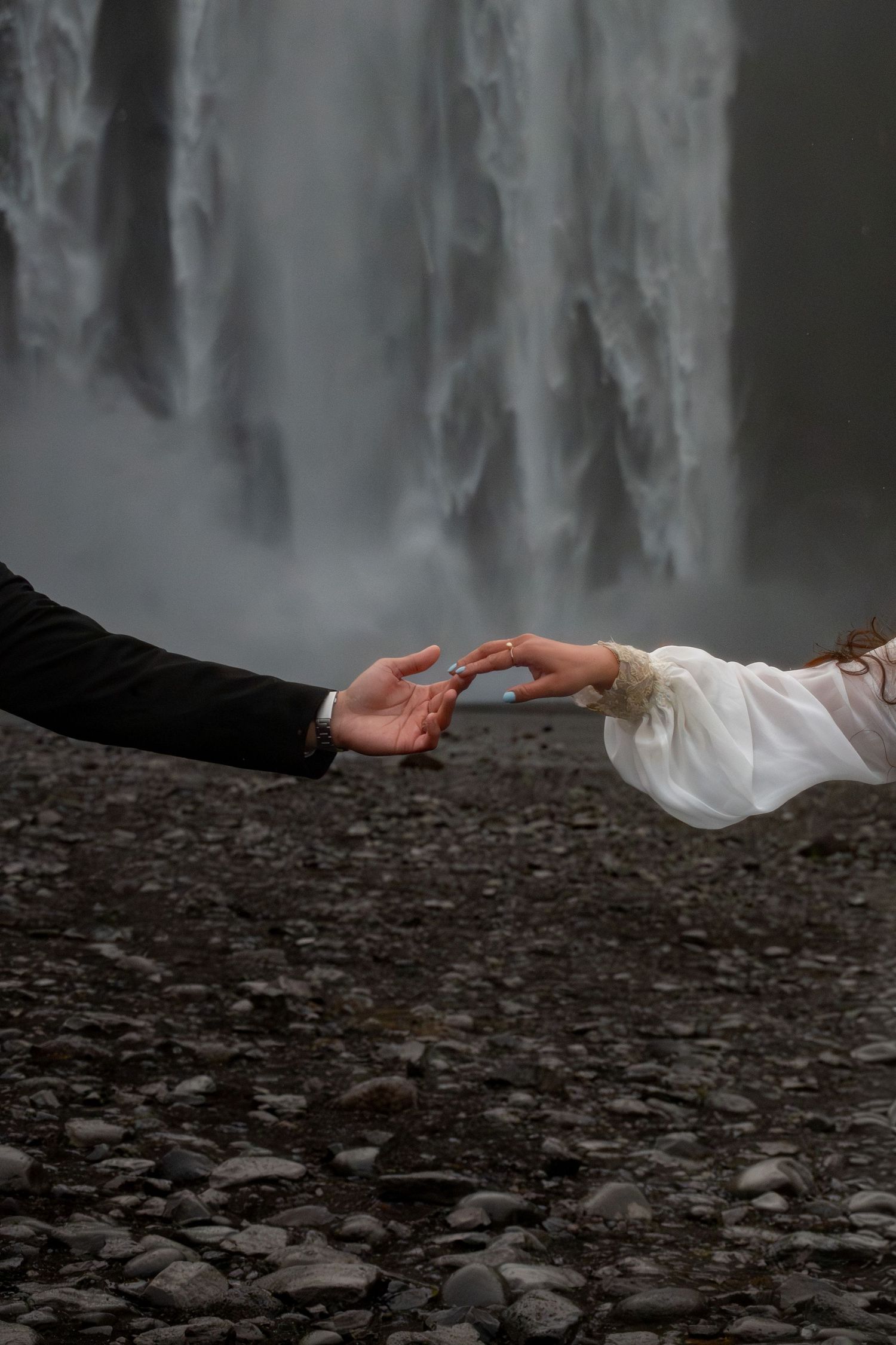 Close-up of bride and groom reaching for each other's hands with Skogafoss waterfall in Iceland behind them.