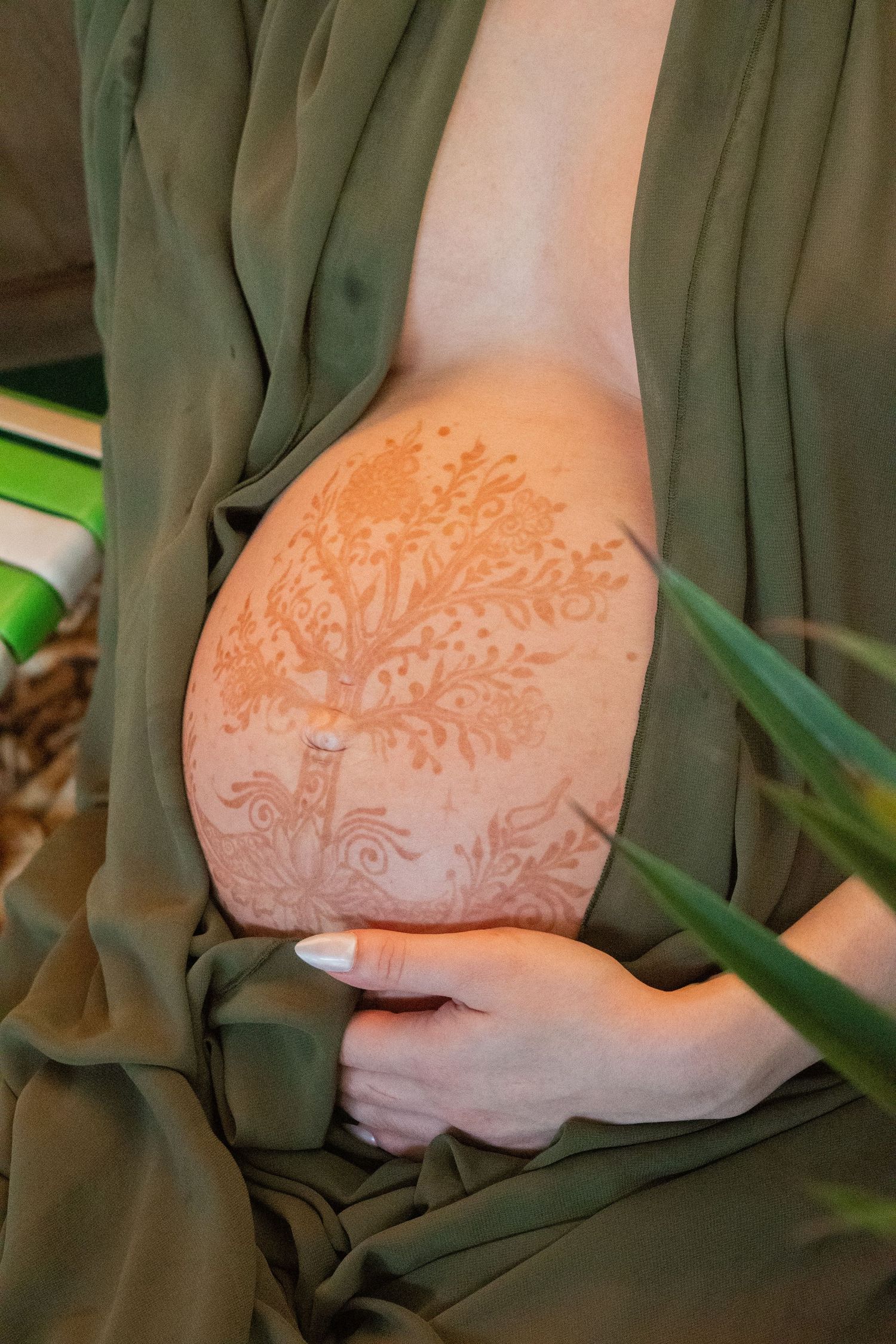 Pregnant woman with intricate henna tree design painted on her baby bump, wearing a green draped dress.