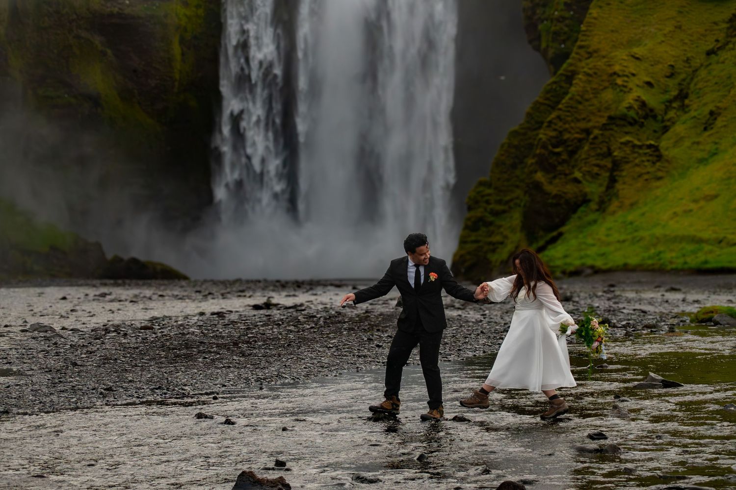 Couple in wedding attire holding hands near Skogafoss waterfall in Iceland.