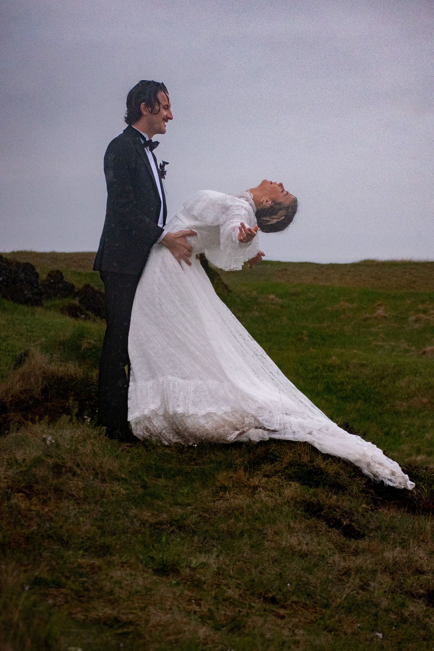 Bride in white gown dips back with groom in tuxedo on grassy hillside during overcast outdoor wedding.