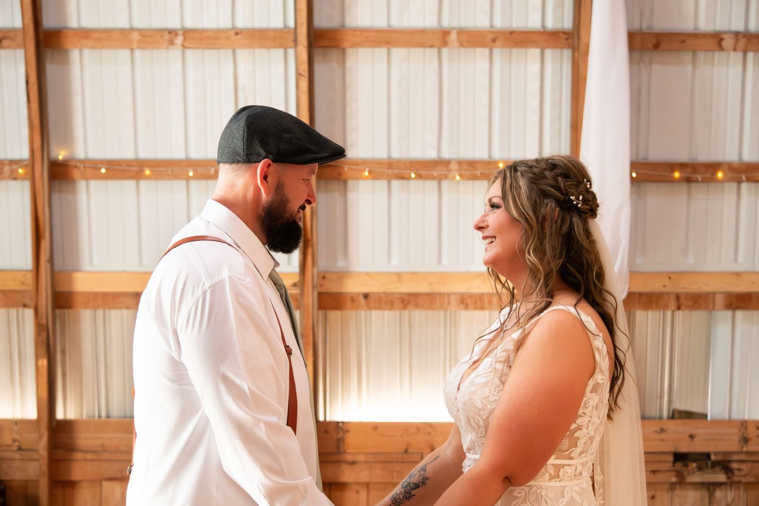 Bride and bearded groom in a flat cap smiling at each other during wedding ceremony in a barn.
