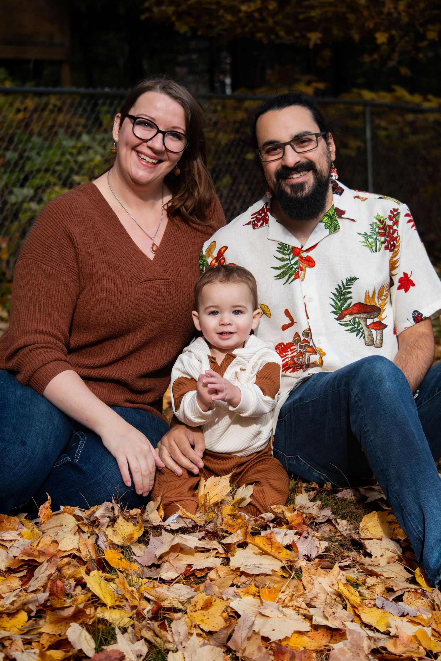 Happy family of three sitting in colorful autumn leaves, parents smiling with baby in fall outdoor portrait.