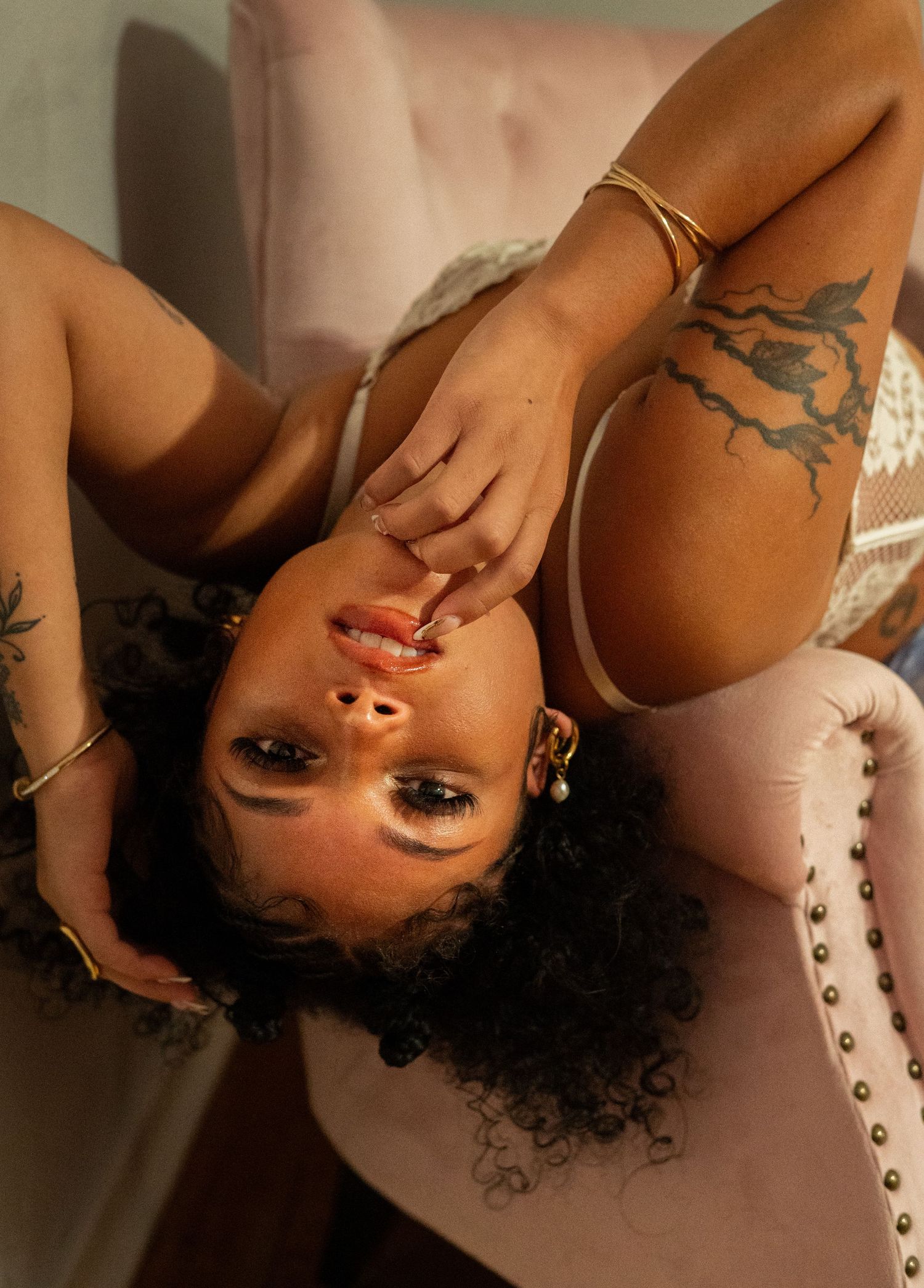 A woman with curly hair and tattoos poses lying back on a pink velvet chair wearing gold jewelry.
