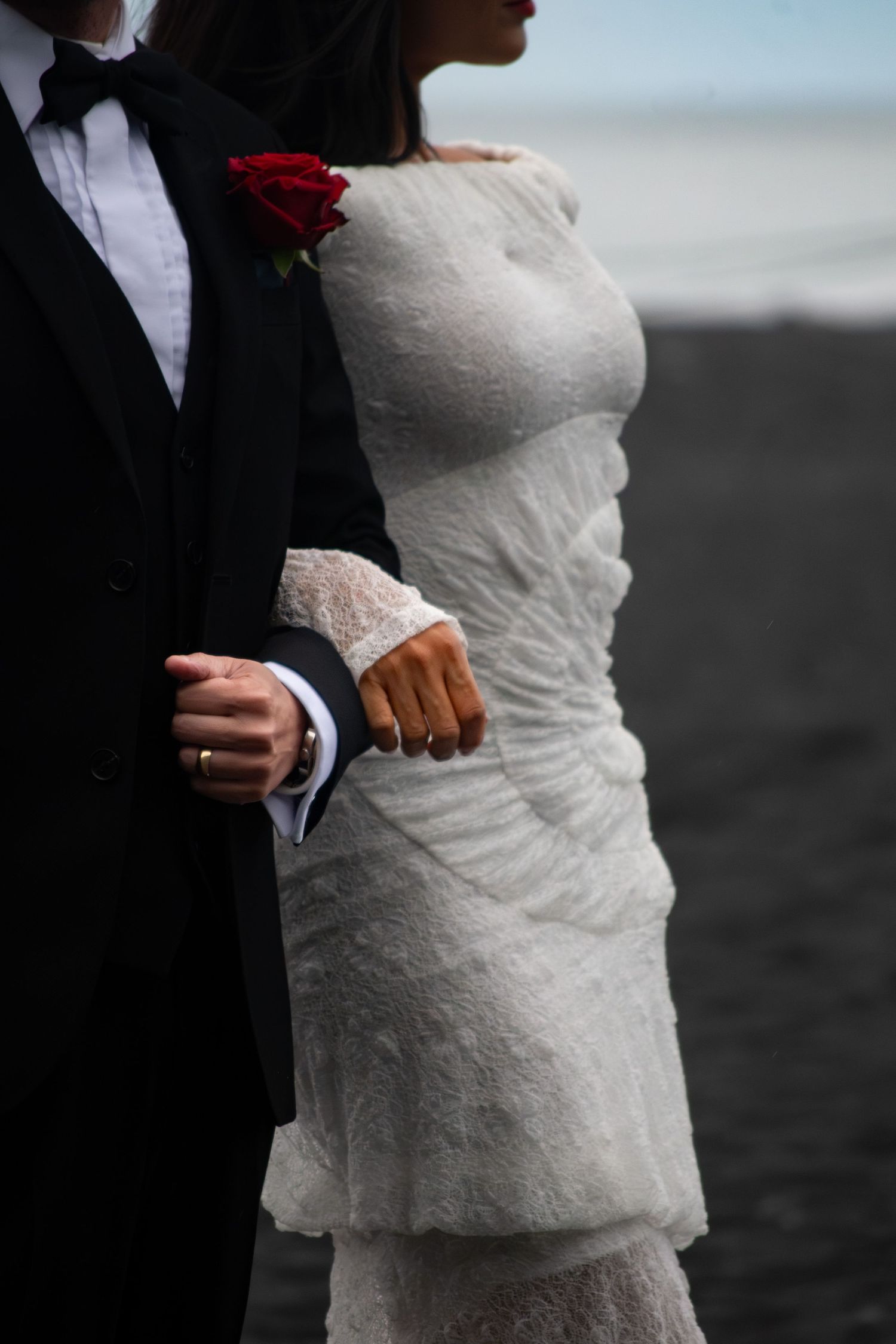 Bride in white lace gown holds groom's arm on black sand beach, red rose boutonniere visible.