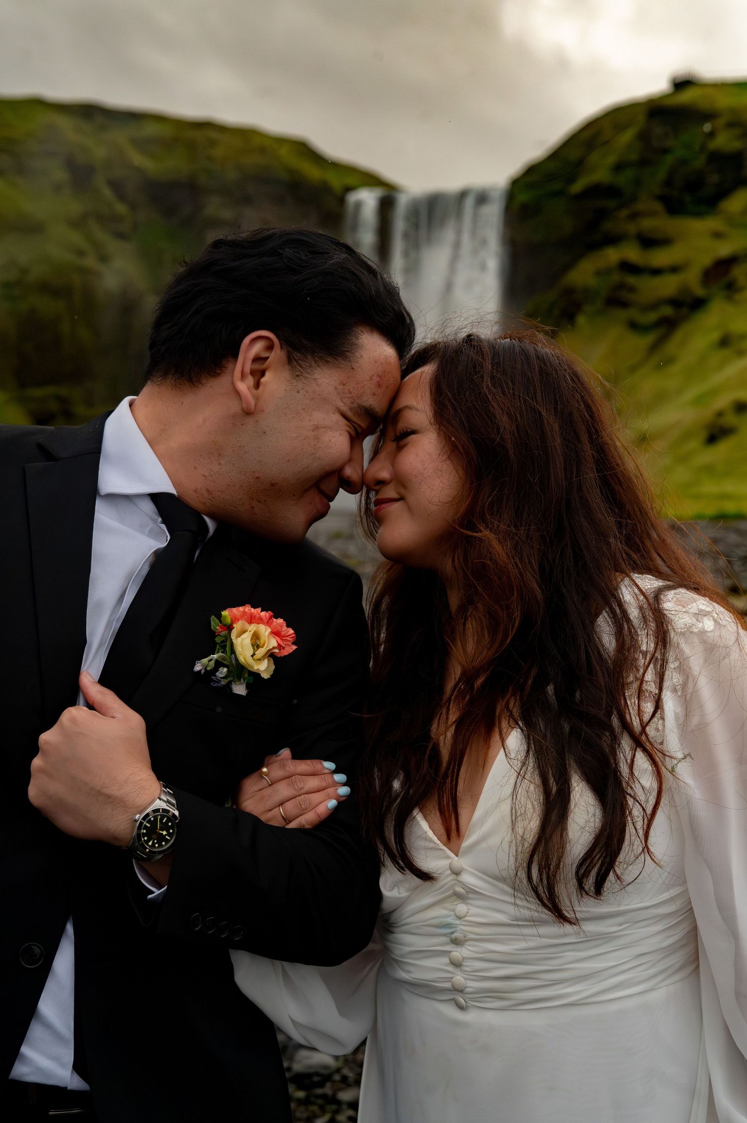Couple in wedding attire sharing a romantic moment with foreheads touching, waterfall and green cliffs in background.