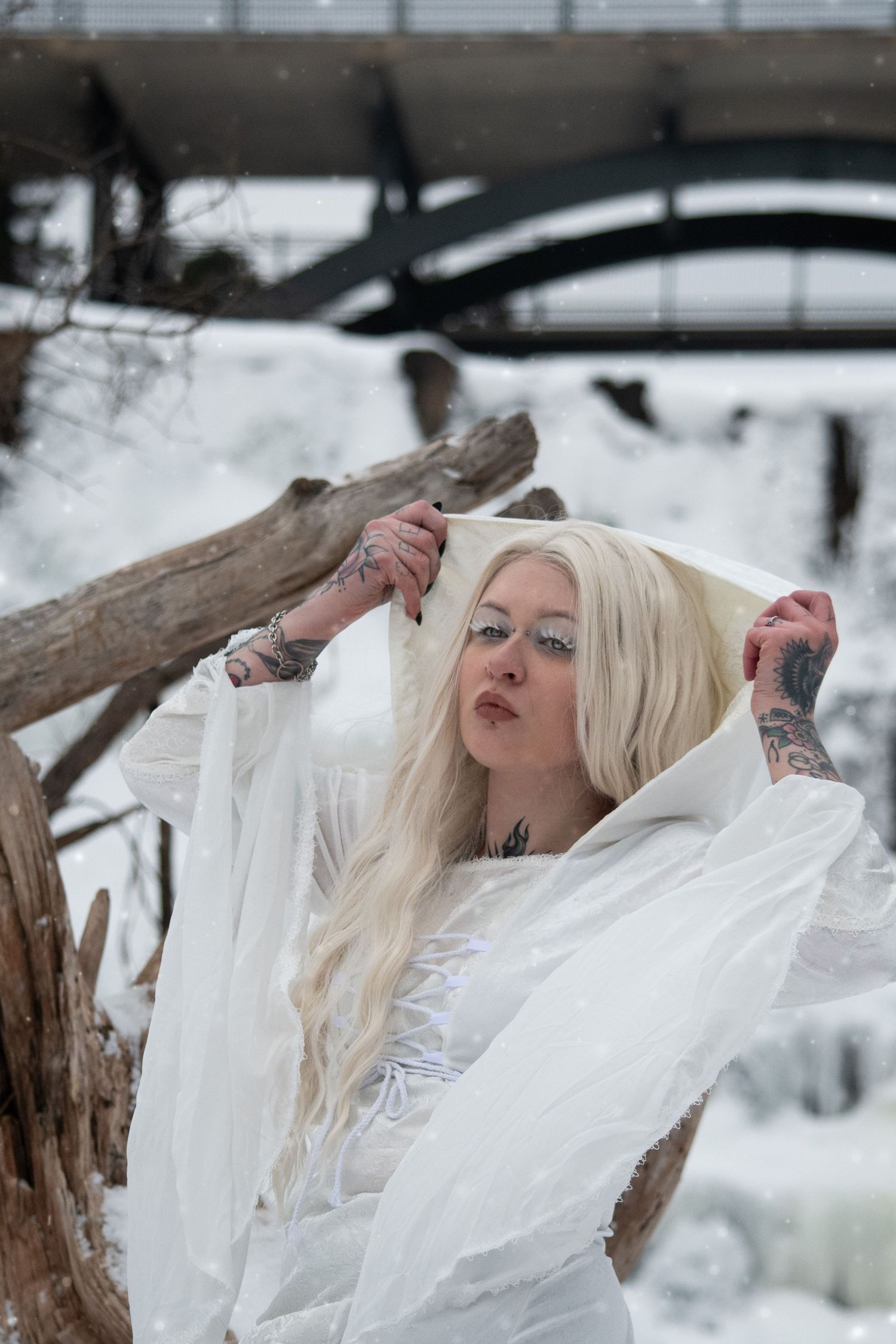 Tattooed blonde woman adjusting white veil near driftwood with bridge in snowy background.