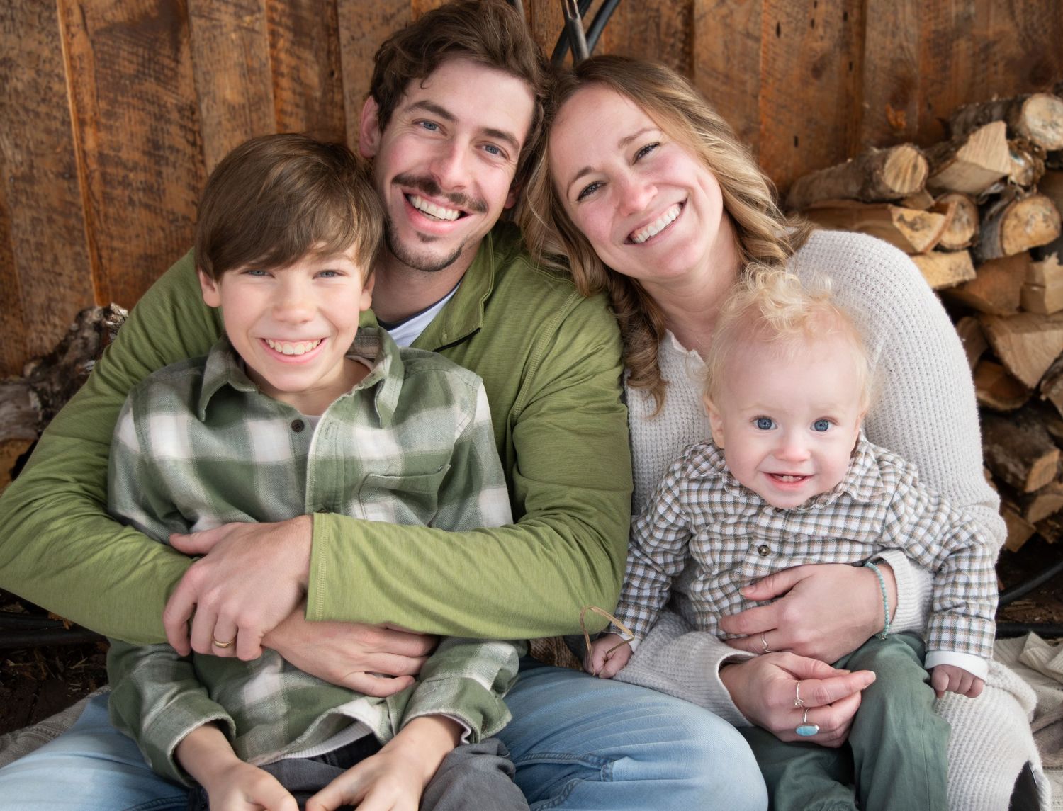 Smiling family of four posing together in front of rustic wooden log wall backdrop.